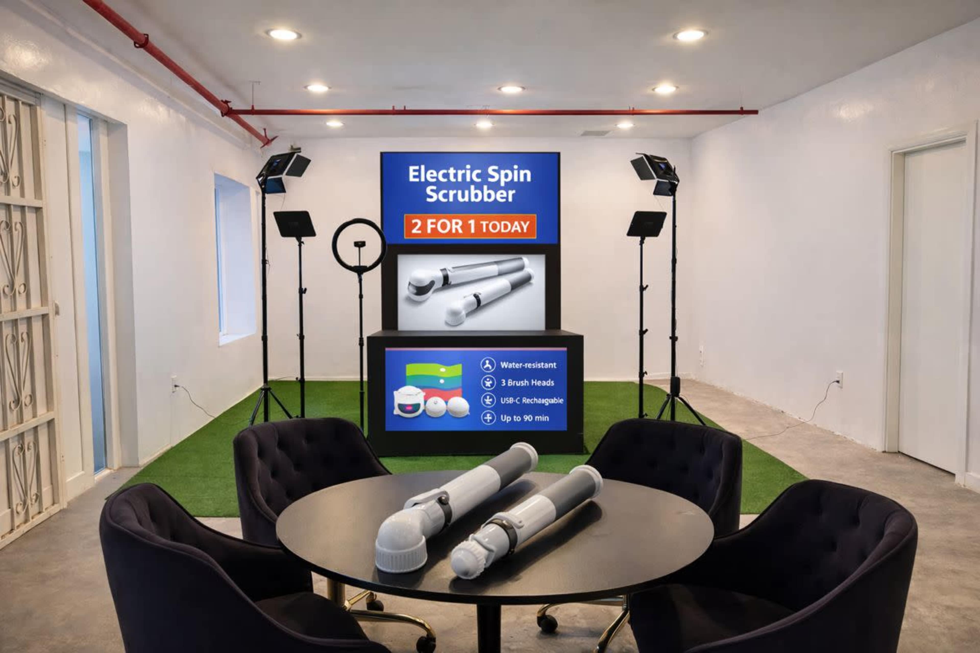 The image shows a modern promotional setup featuring a display for an electric spin scrubber, positioned in a well-lit room with a circular table and four black chairs.