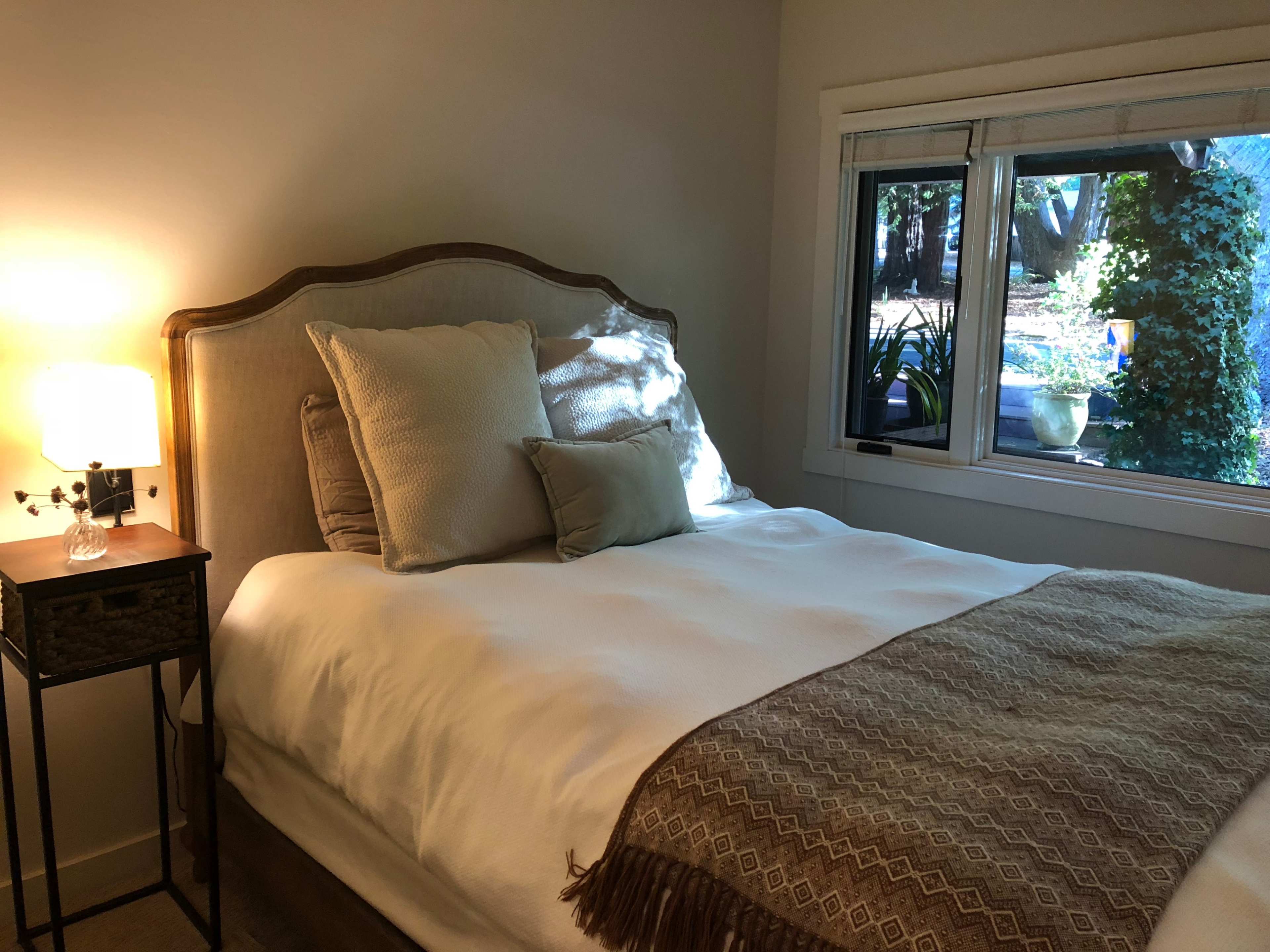 A softly lit bedroom with a bed adorned with pillows, a throw blanket, and a bedside table featuring a lamp beside a window with views of greenery outside.