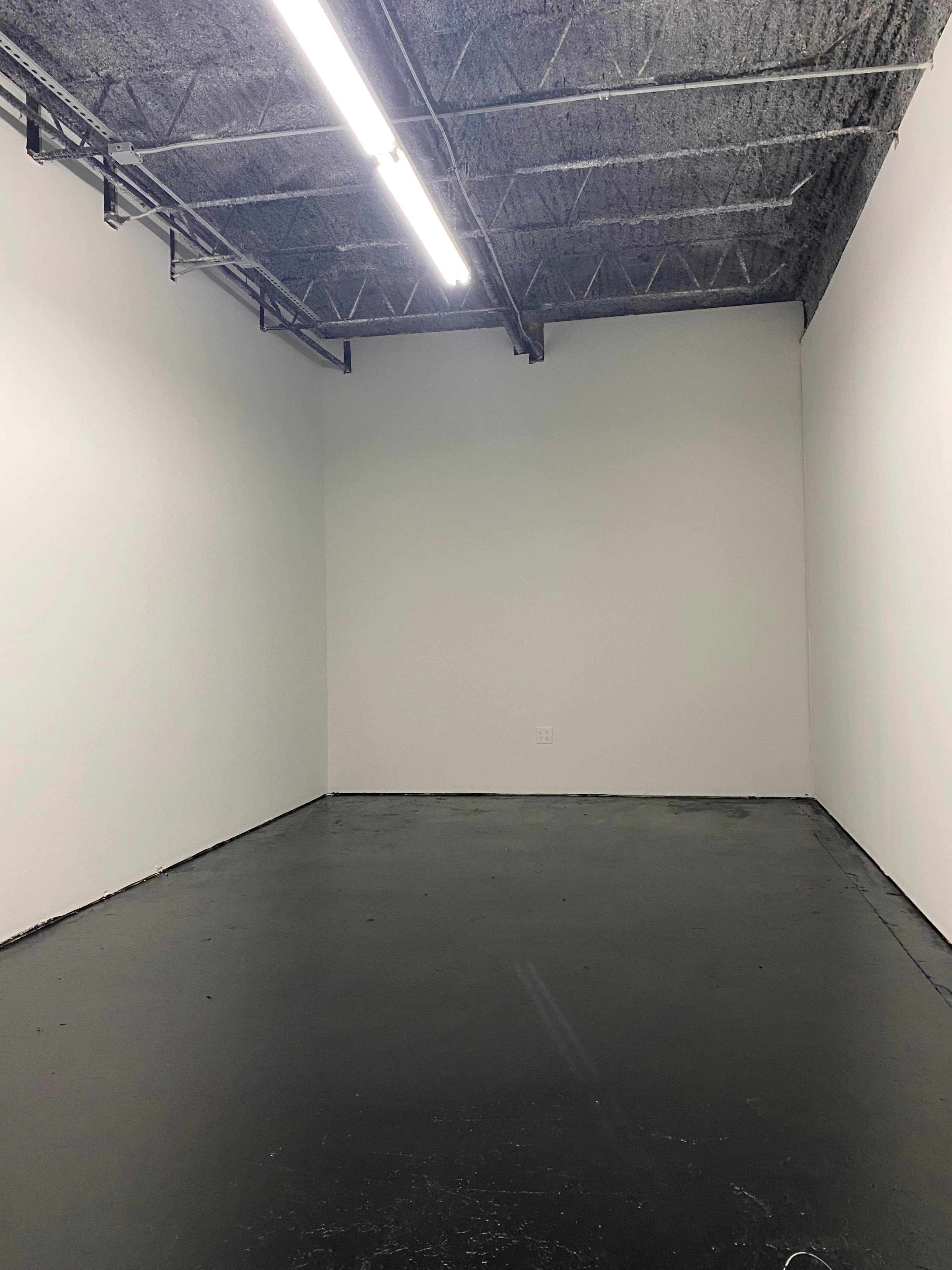 A bare, empty room with a concrete floor and white walls, illuminated by fluorescent lights from the ceiling.