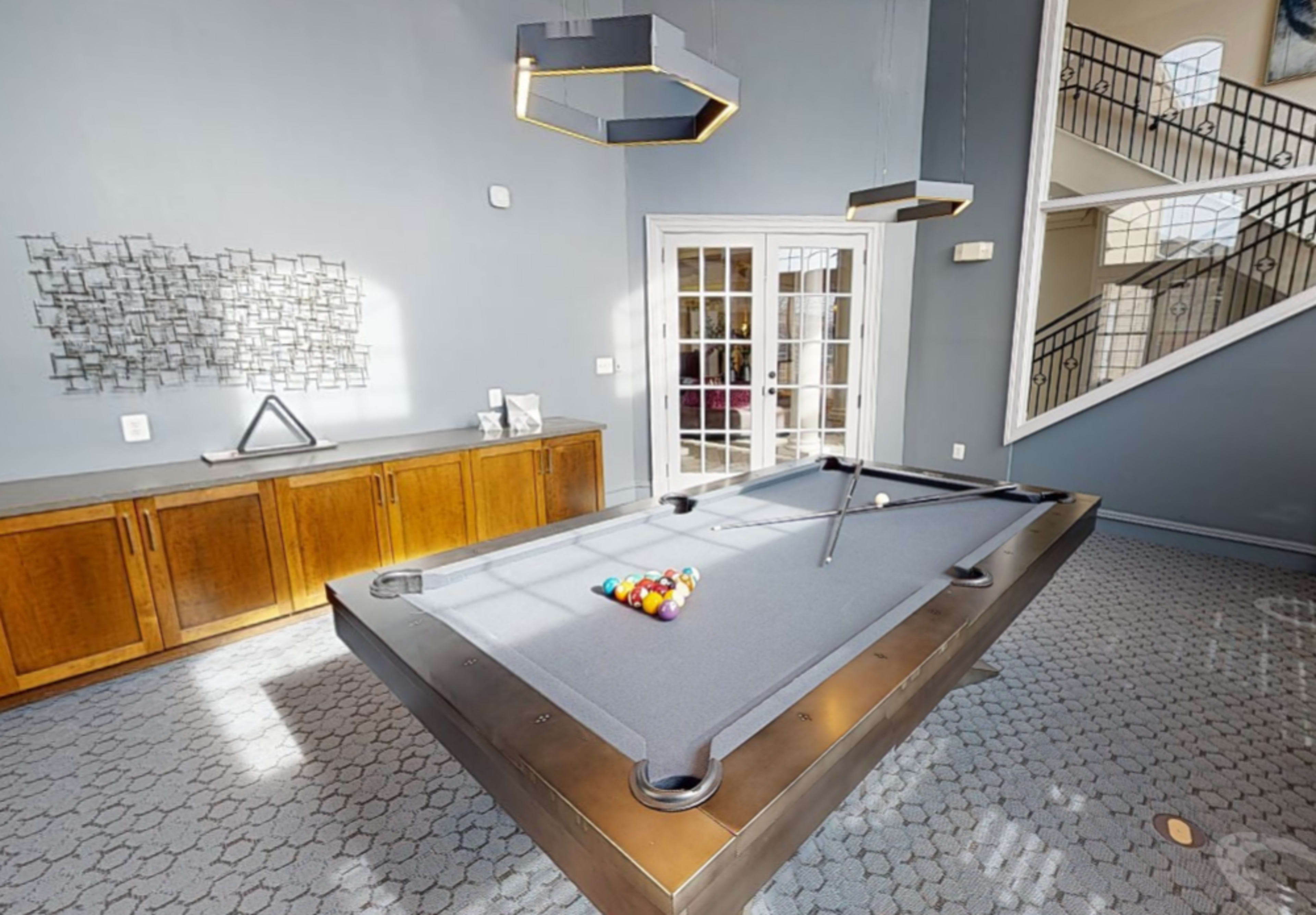 A game room with a pool table, gray carpet, modern lighting fixtures, and large windows leading to a staircase.