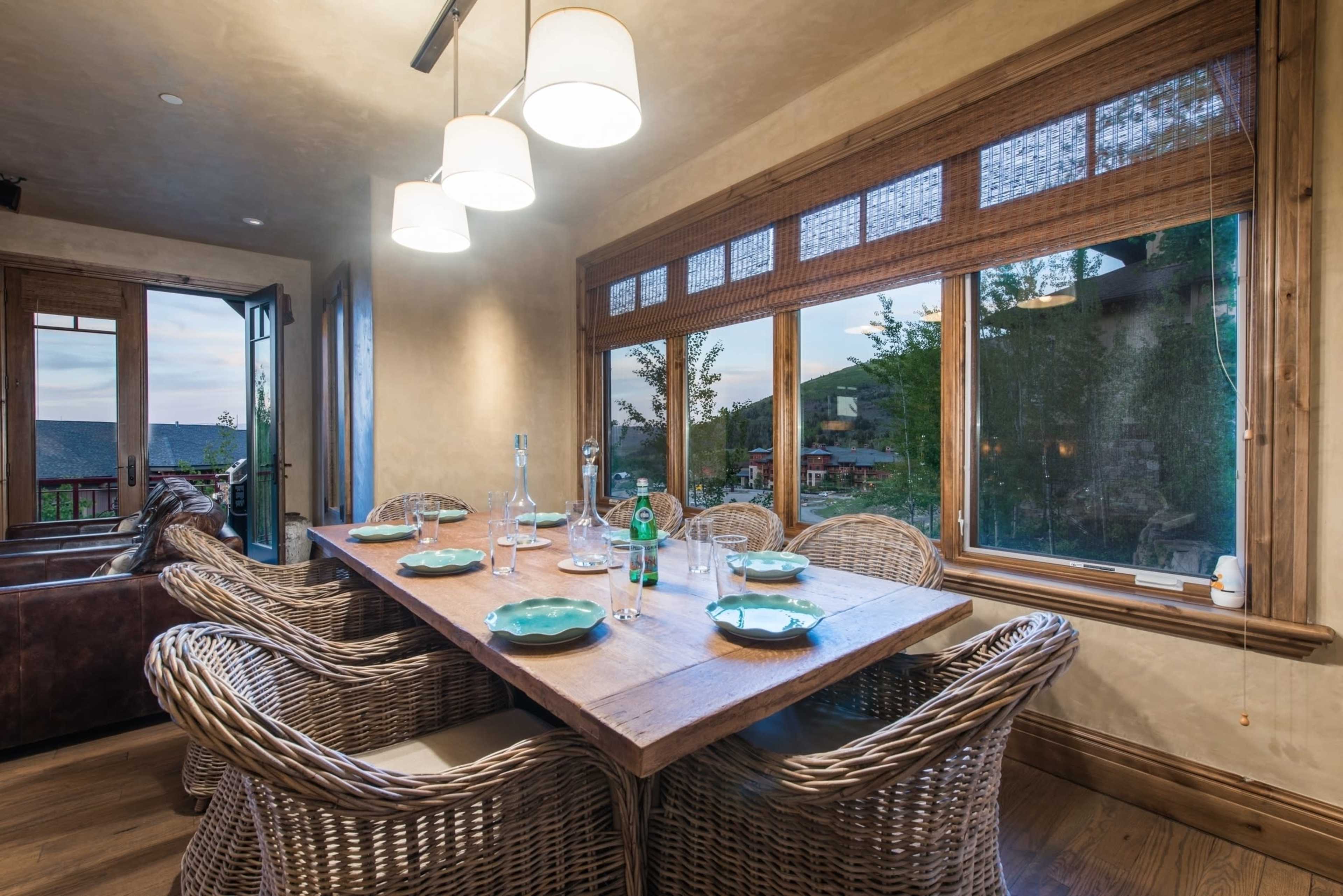A wooden dining table with plates and glasses is set in a room with large windows and woven chairs.