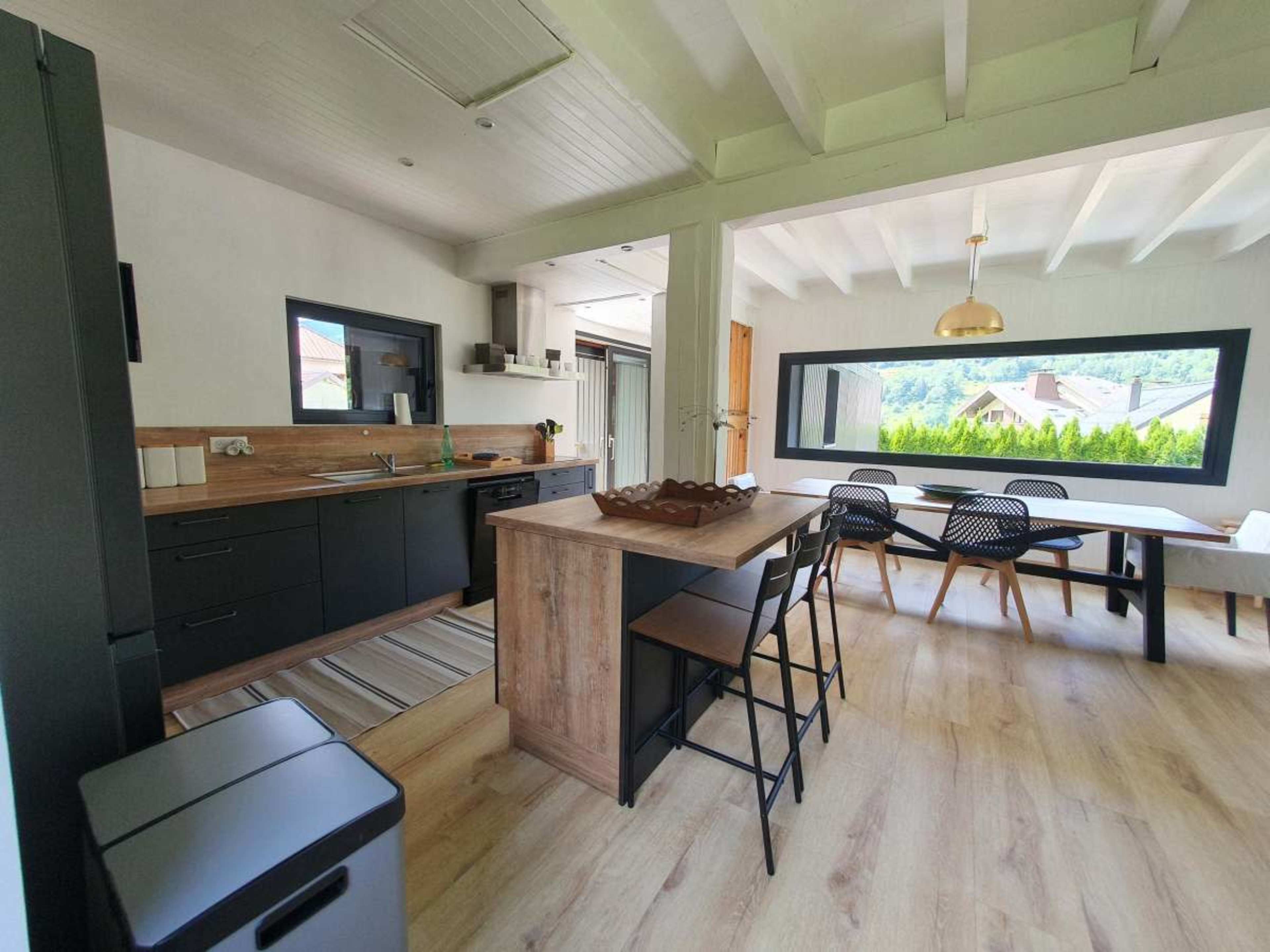 The image shows a modern kitchen and dining area featuring black cabinets, a wooden island, and large windows with a view of greenery outside.