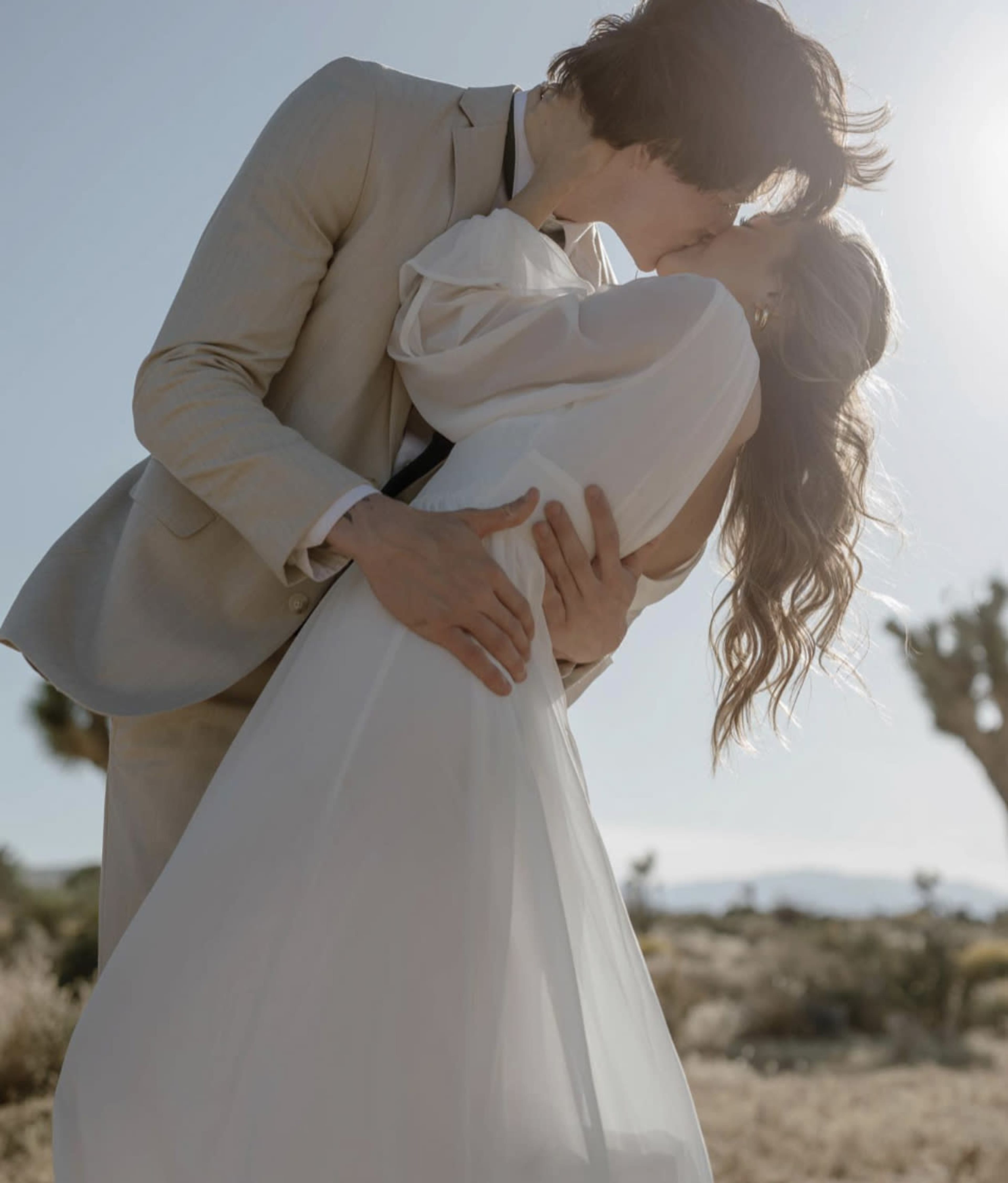 A couple kisses each other in a desert landscape while the sun shines overhead.