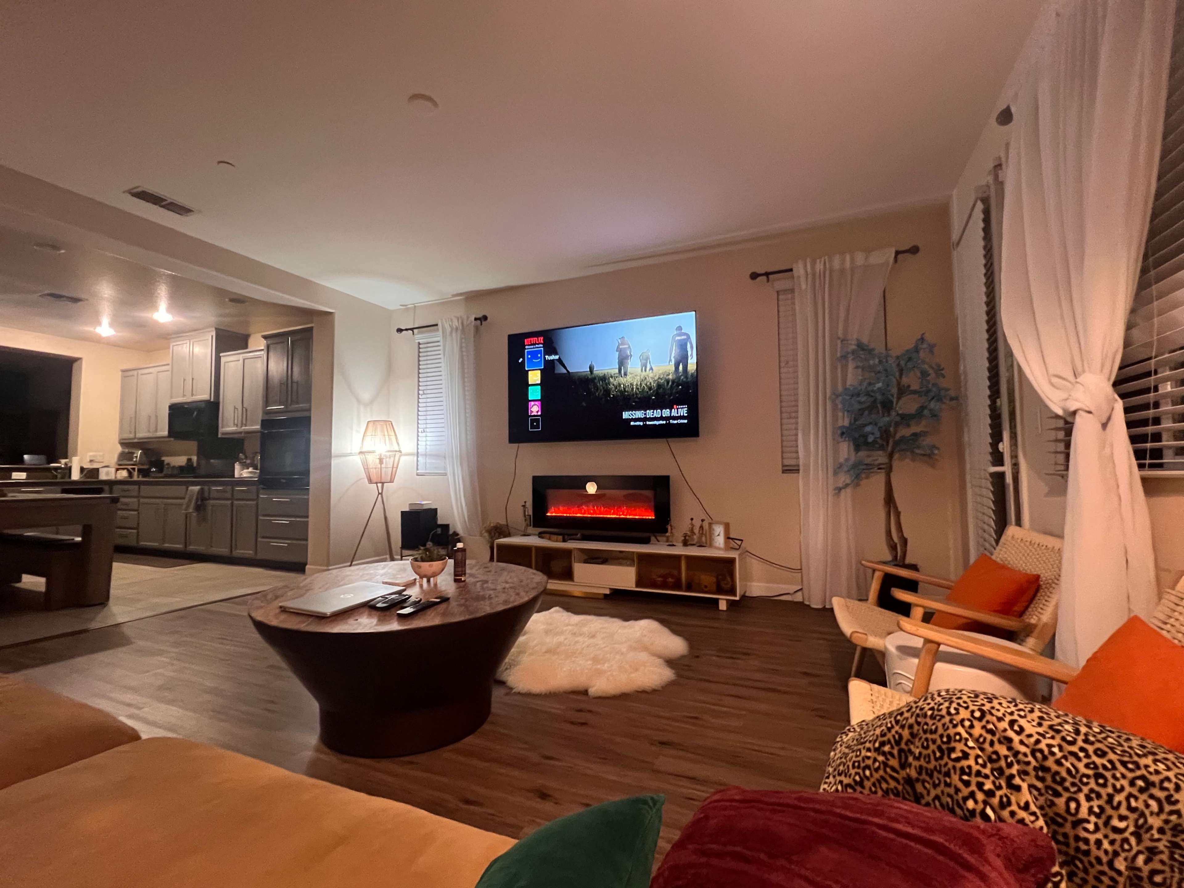 A cozy living room features a large television mounted on the wall, a round coffee table, and a sectional sofa with colorful cushions, while a kitchen area is visible in the background.