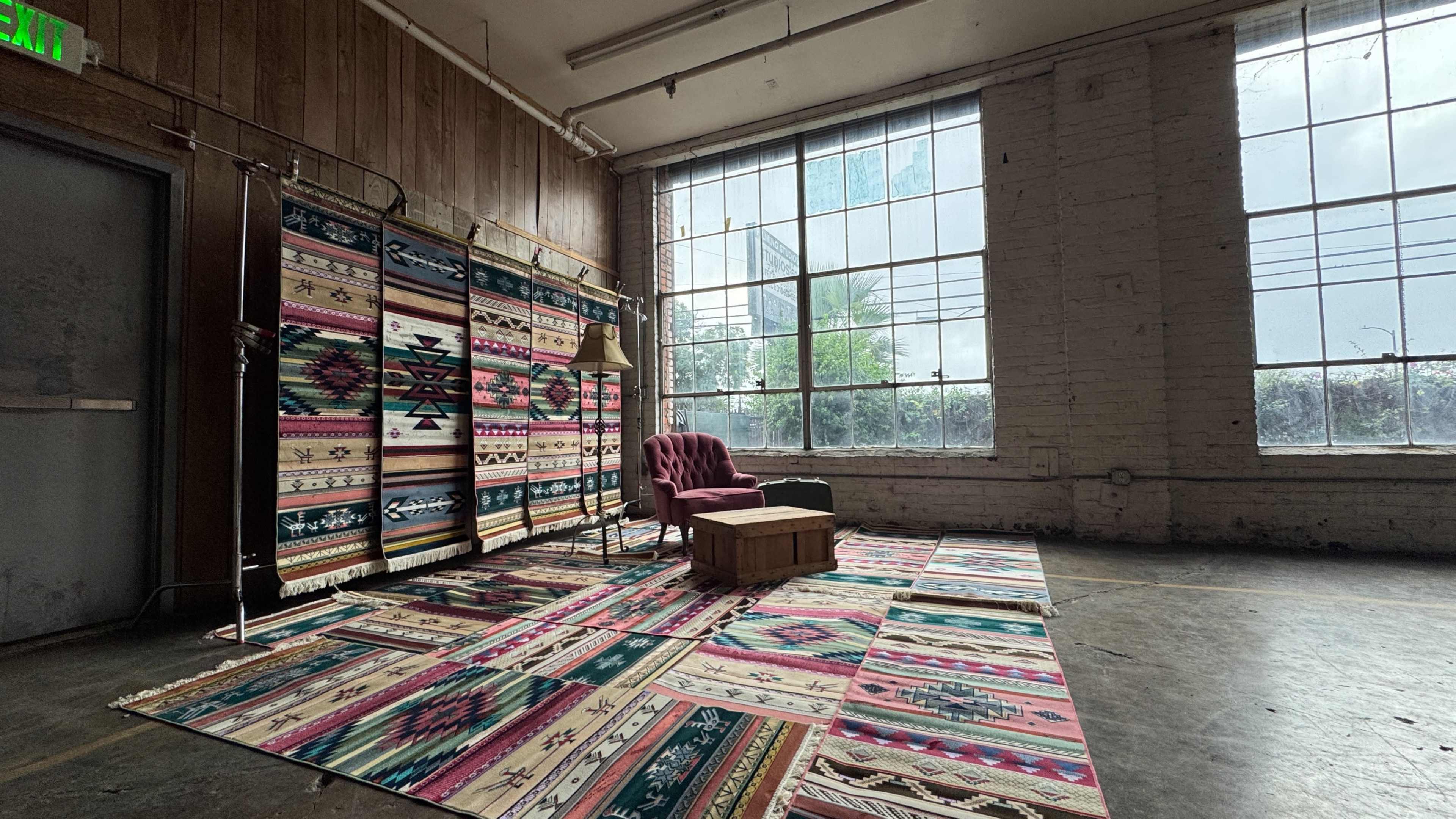 The image shows a spacious room with large windows, featuring a colorful patterned area rug, a wooden coffee table, a purple chair, and woven wall hangings.