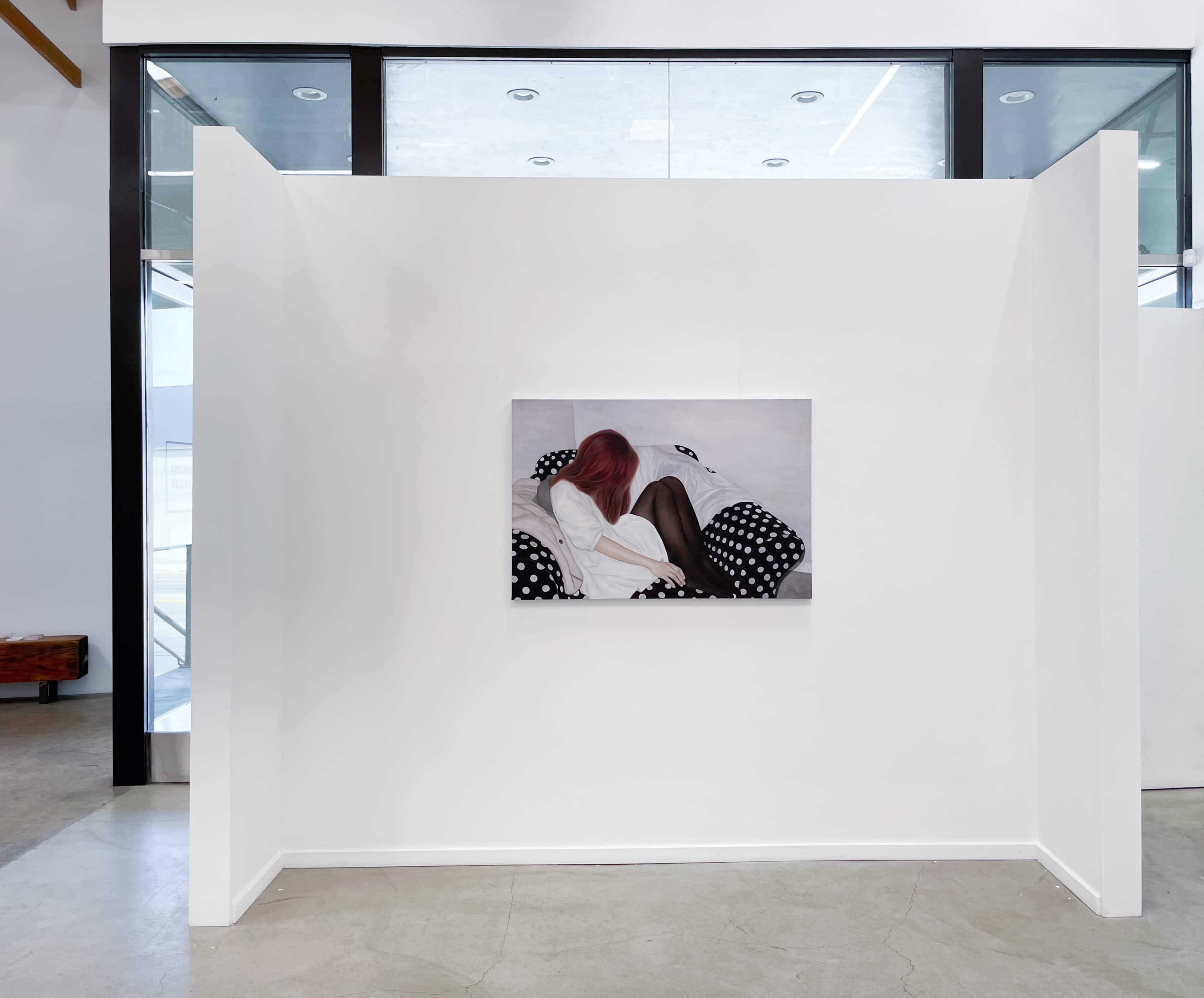 The image shows a minimalist gallery space with a large artwork featuring a figure with pink hair against a white wall, framed by two white partitions.