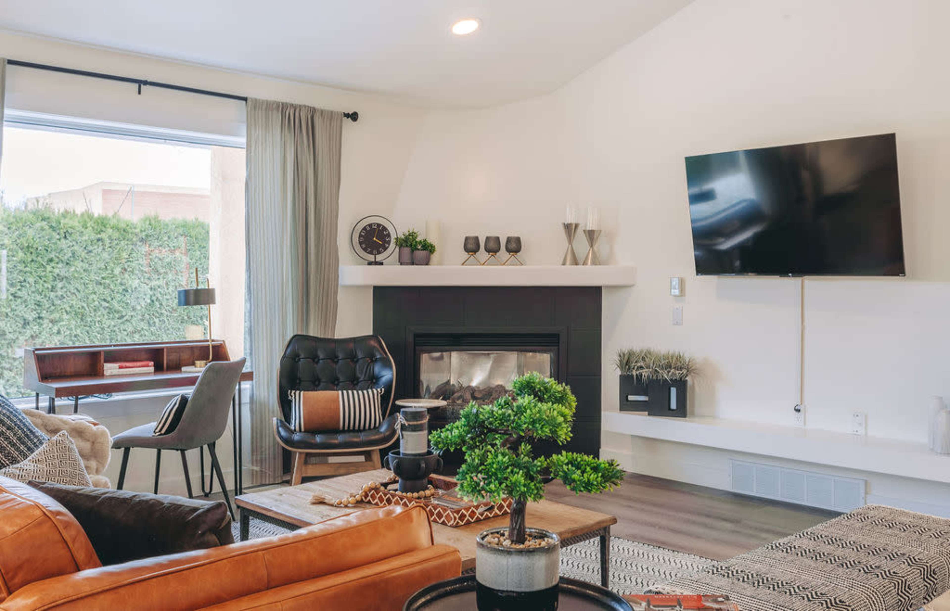 The image shows a modern living room featuring a fireplace, a television mounted on the wall, and a mix of seating options including a leather sofa and a black armchair, with a window providing natural light.