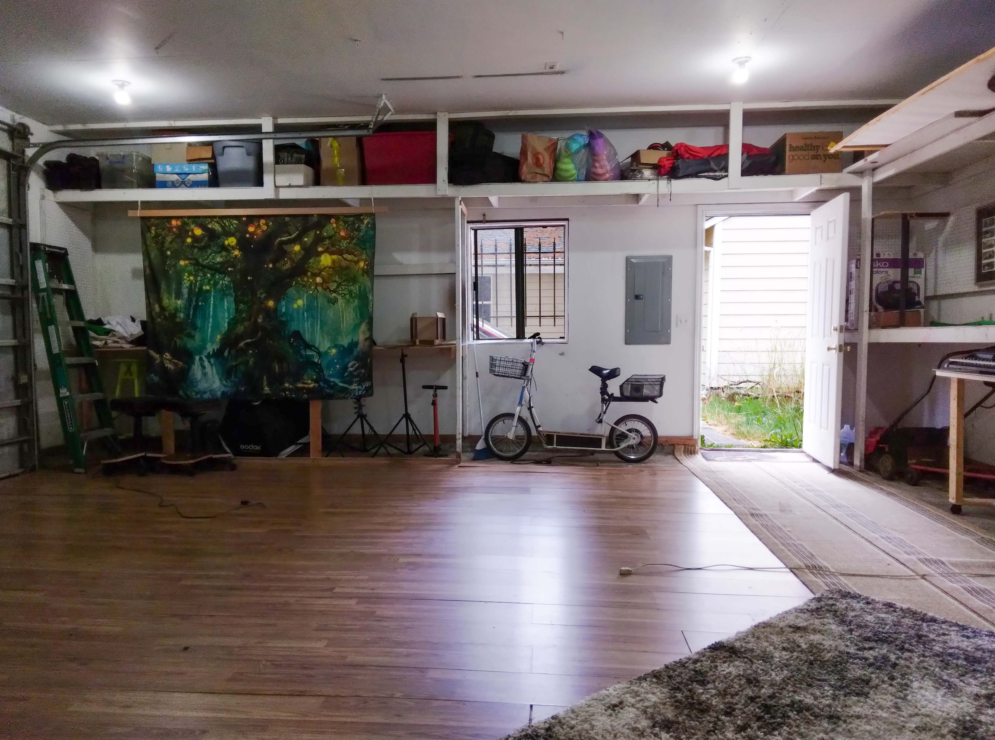 A spacious garage interior with wooden flooring, a large tree painting on an easel, a bicycle, and shelving filled with various items.