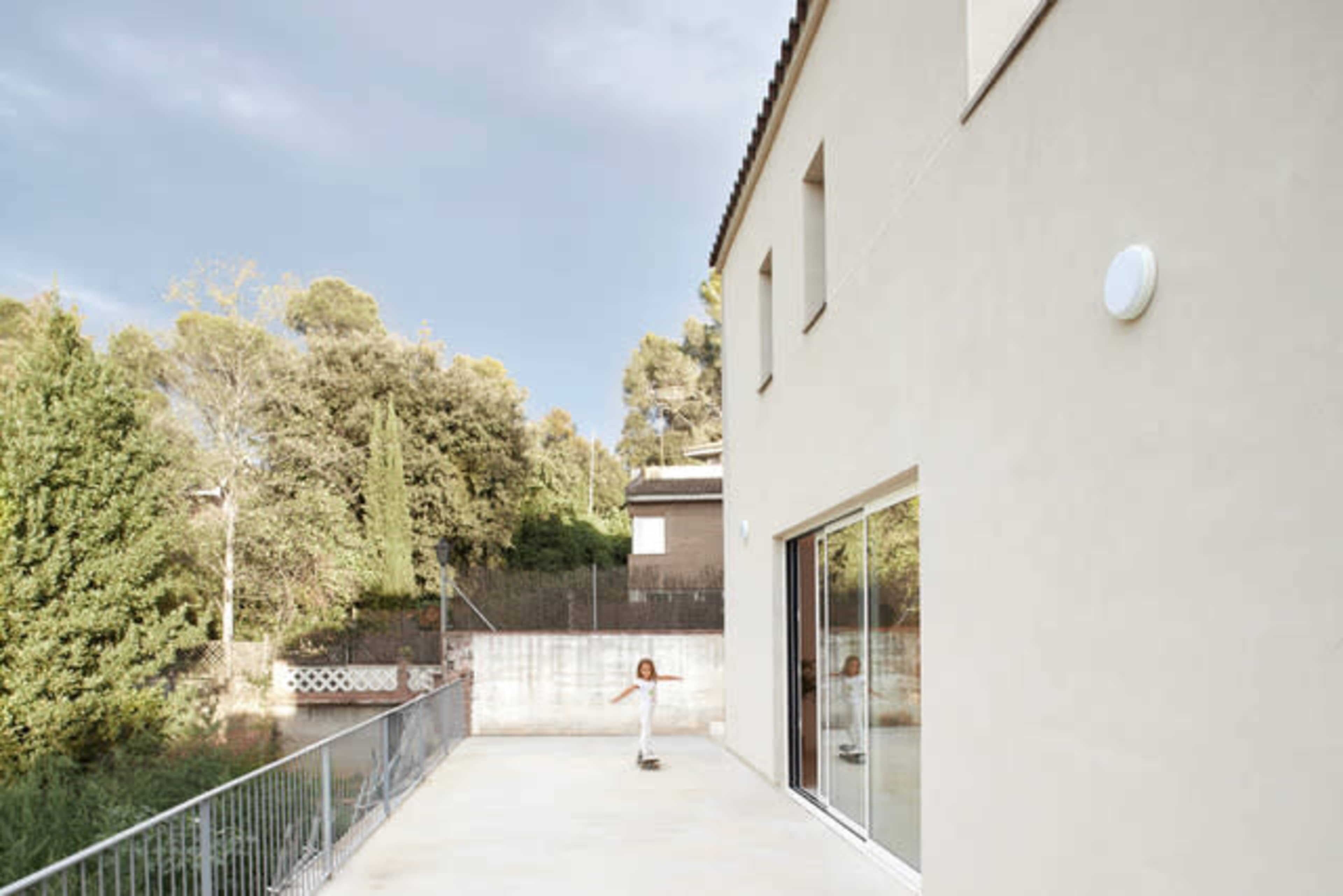 A child skateboards on a smooth patio beside a modern house, with trees and another building visible in the background.