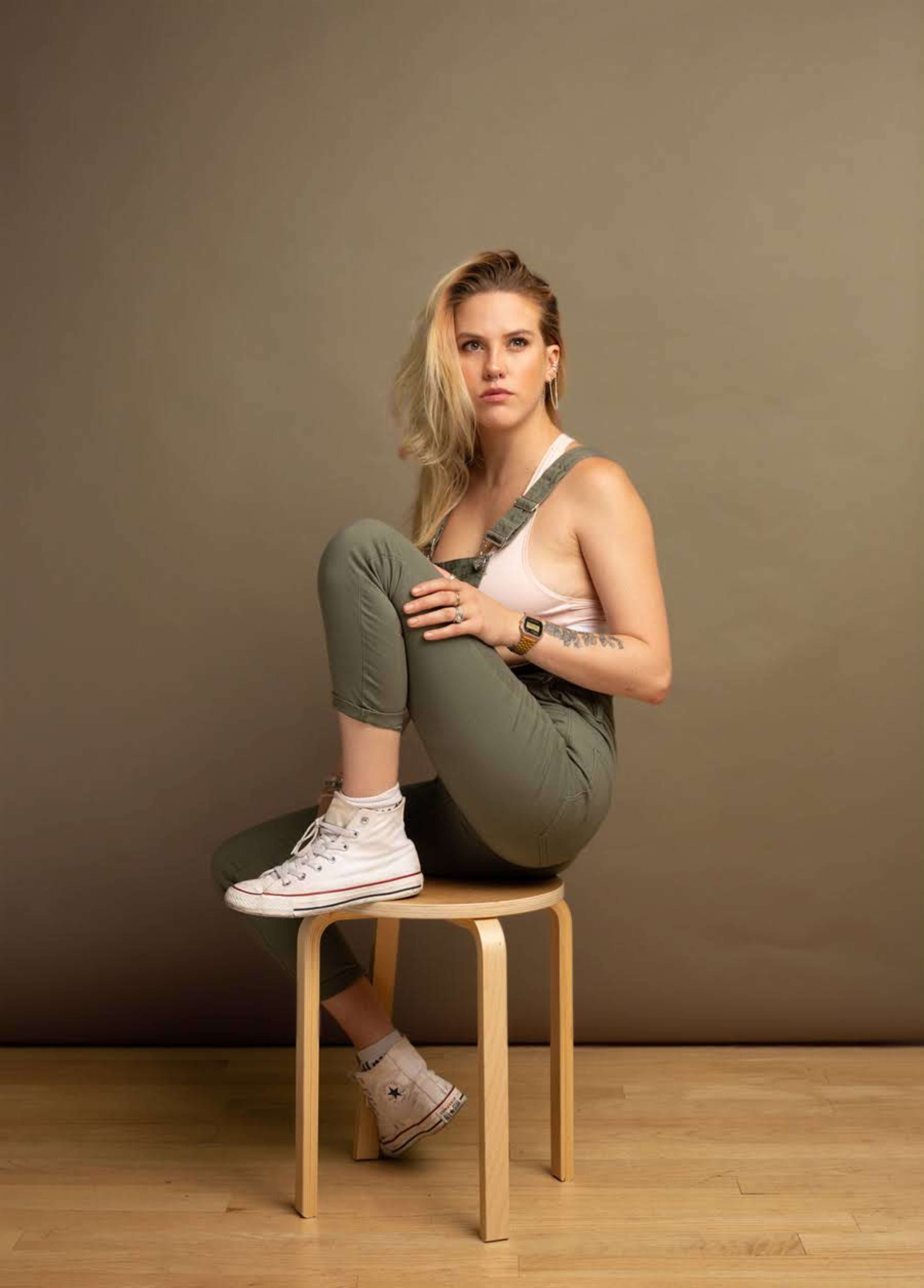 A person with blonde hair sits on a wooden stool with one leg folded beneath them and the other bent, wearing a tank top and olive-green pants.
