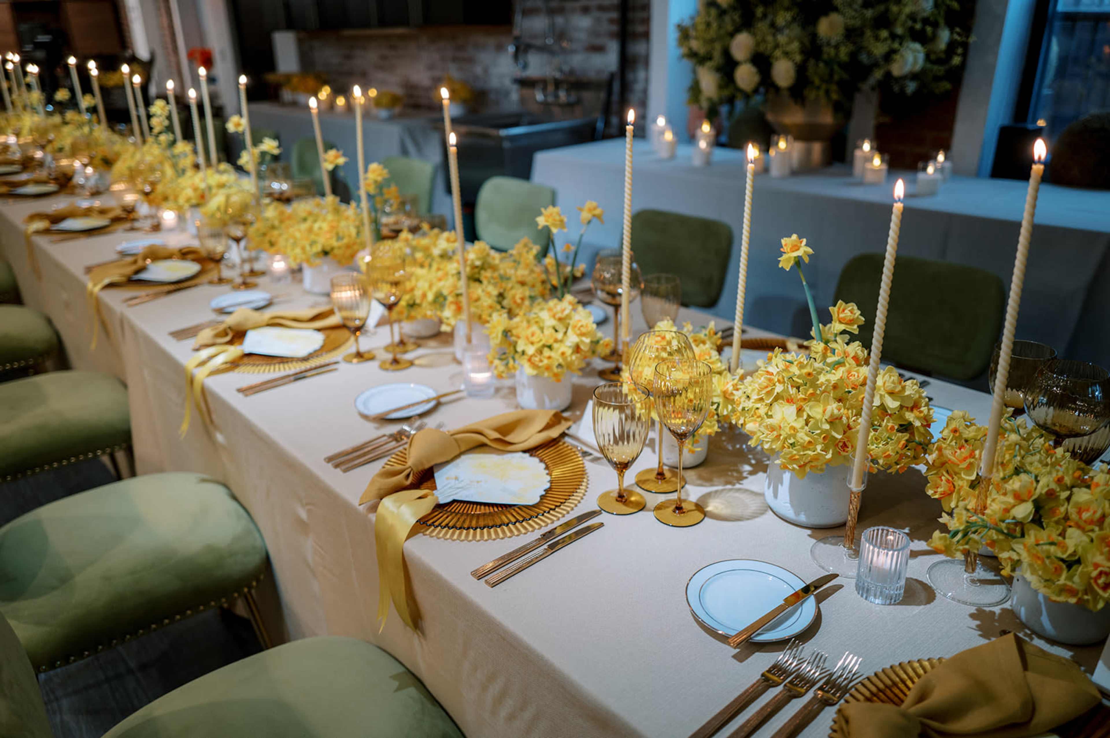 A long dining table is set with yellow flower arrangements, candles, and neatly arranged tableware for a formal gathering.
