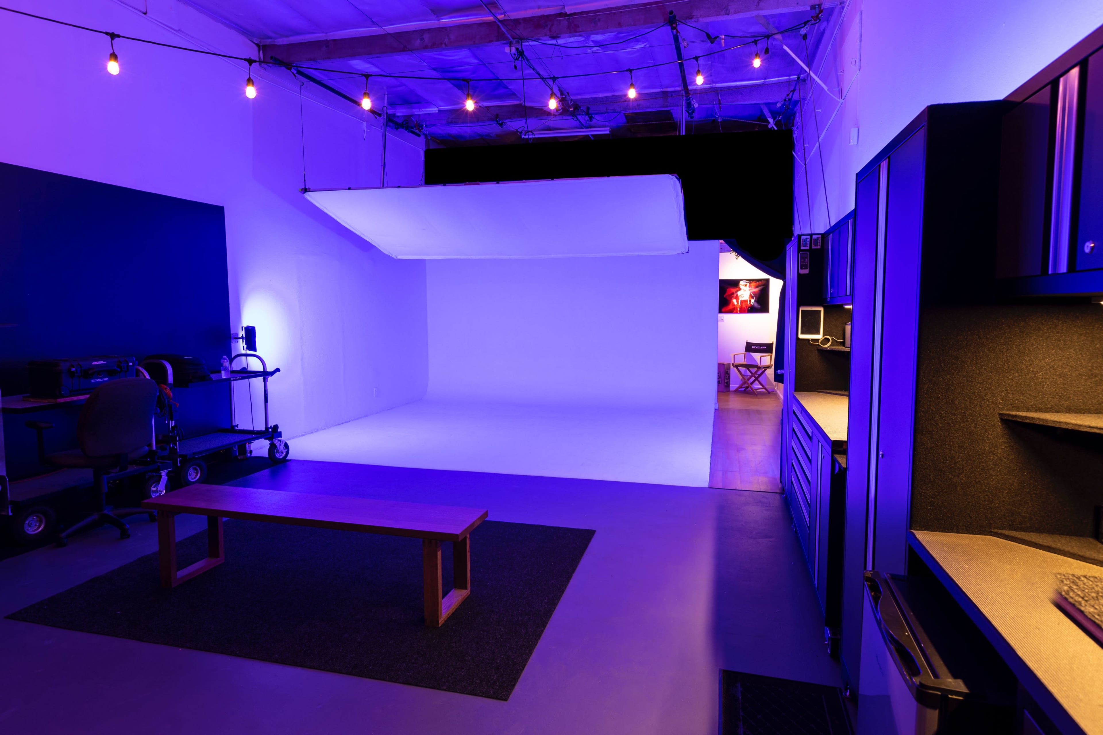 A lit studio space featuring a seamless white backdrop, a wooden bench, and a kitchenette area with overhead lights.