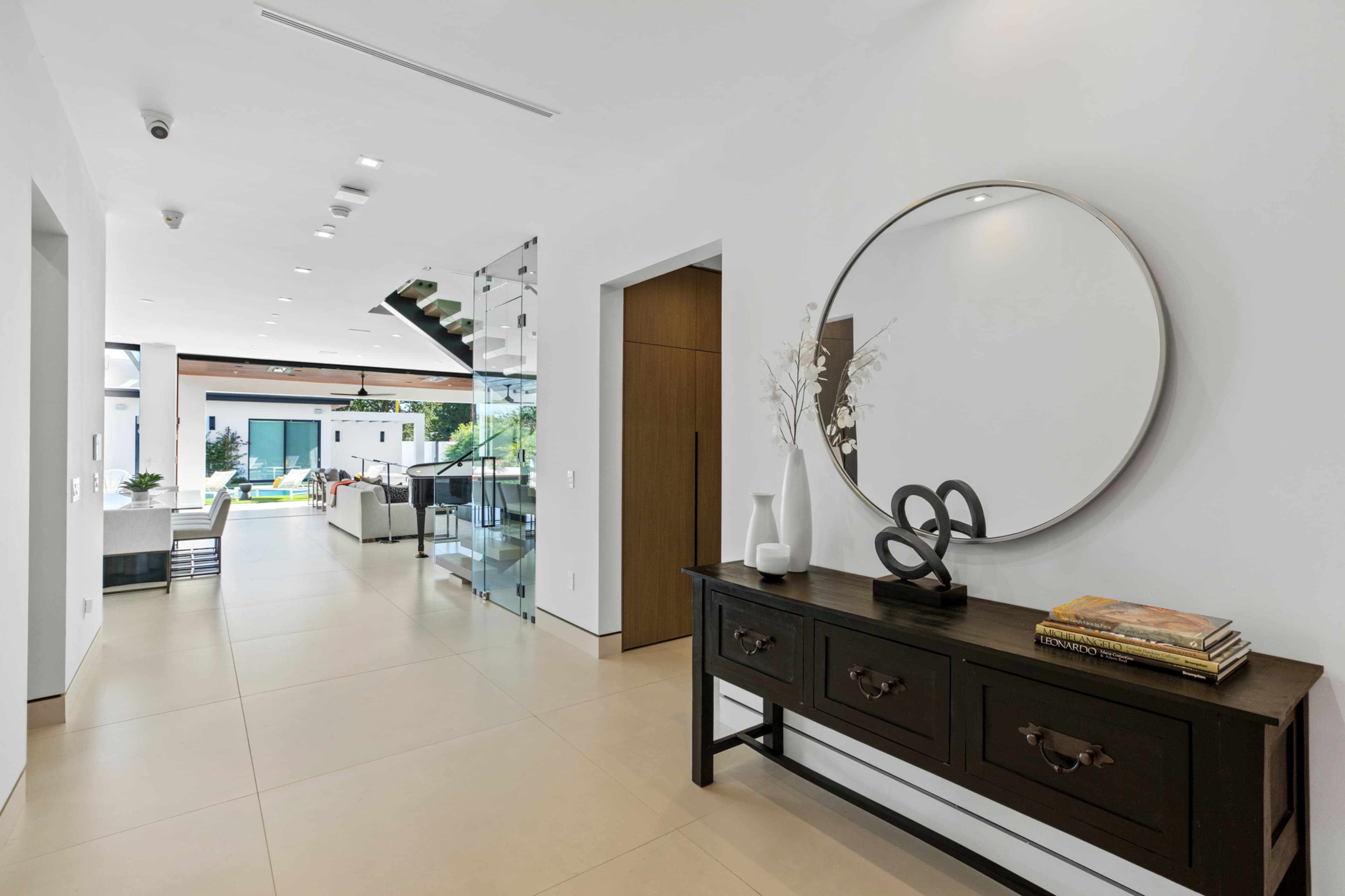 A modern hallway featuring a sleek console table with decor and a round mirror, leading to a spacious living area with large windows.