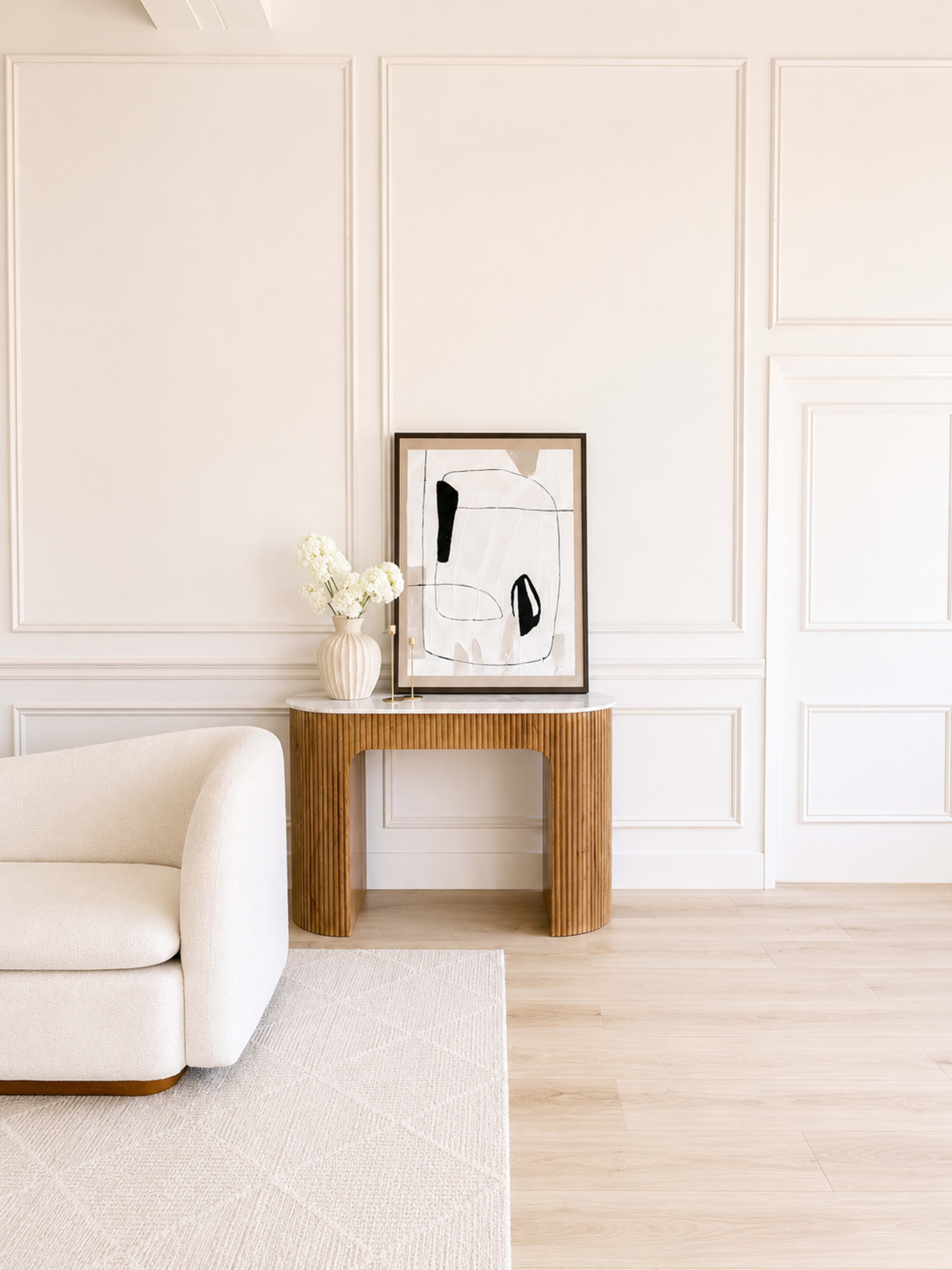 A minimalist living space features a curved, cream-colored sofa next to a wooden table with an abstract painting and a vase of white flowers against a light-colored wall.