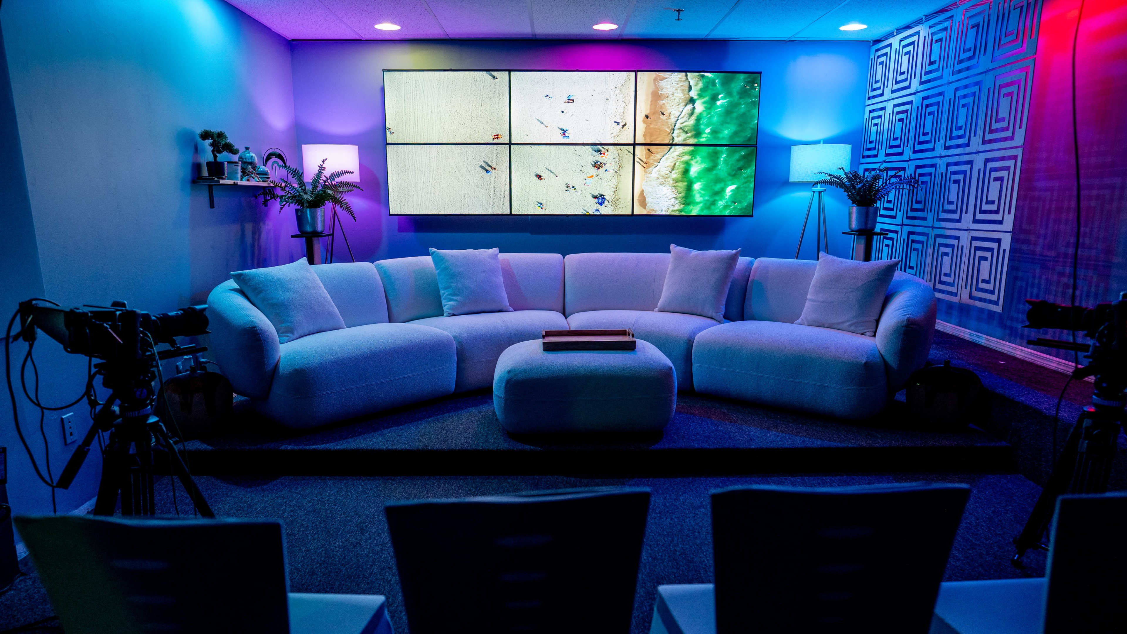 A modern lounge area features a white sectional sofa, a central wooden coffee table, and multiple cameras aimed at a large screen displaying a beach scene.