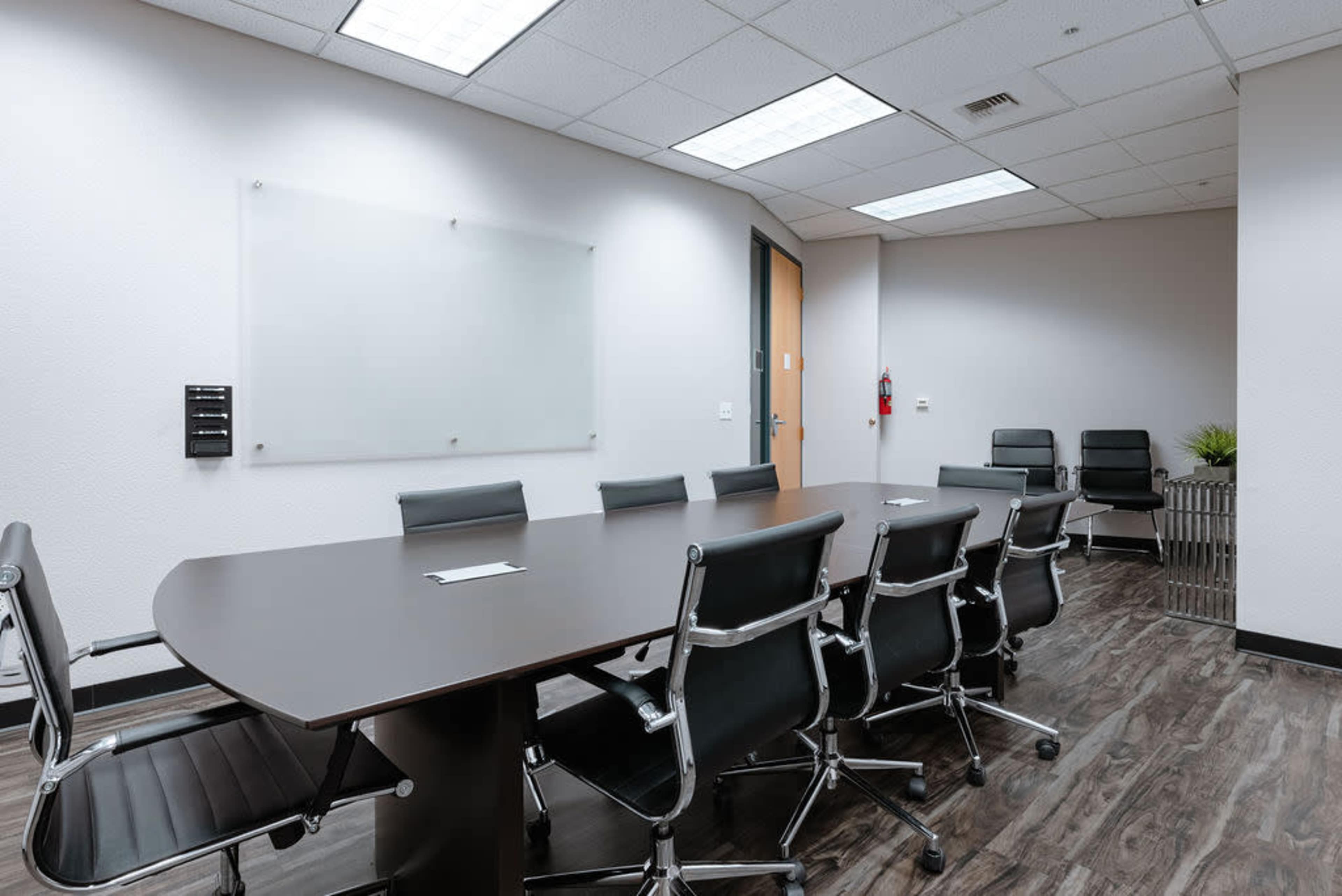 A modern conference room features a long table surrounded by ergonomic chairs and a glass whiteboard on the wall.