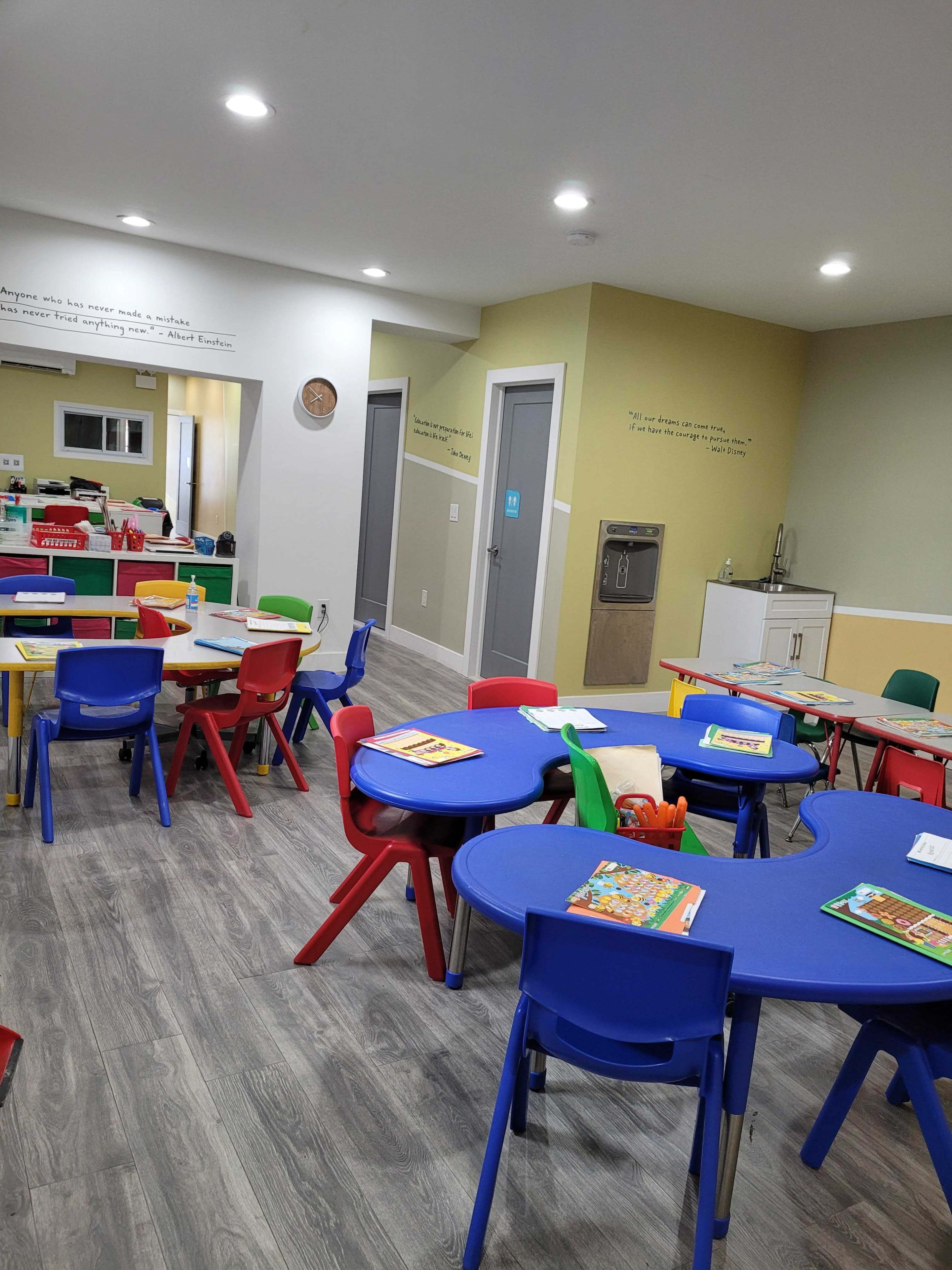 A colorful classroom with several small, round tables and chairs, along with educational materials placed on the tables and a kitchenette area in the background.