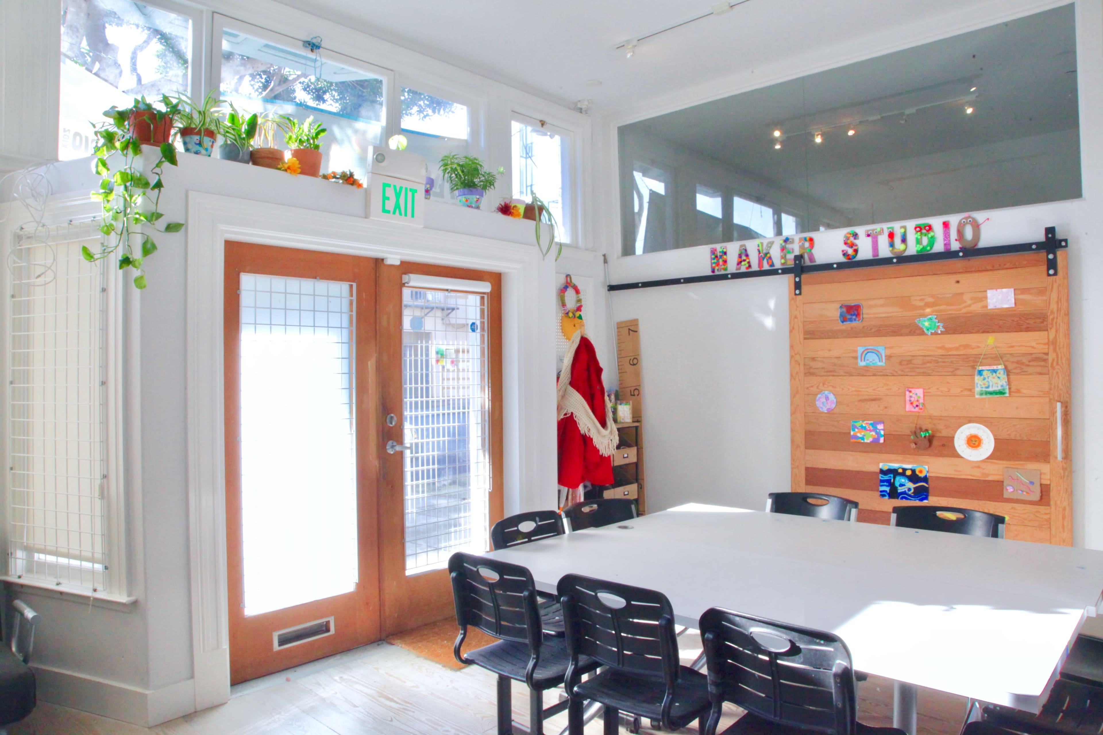 A bright, modern room features a large table surrounded by black chairs, with a door leading outside and a wall decorated with art and plants.
