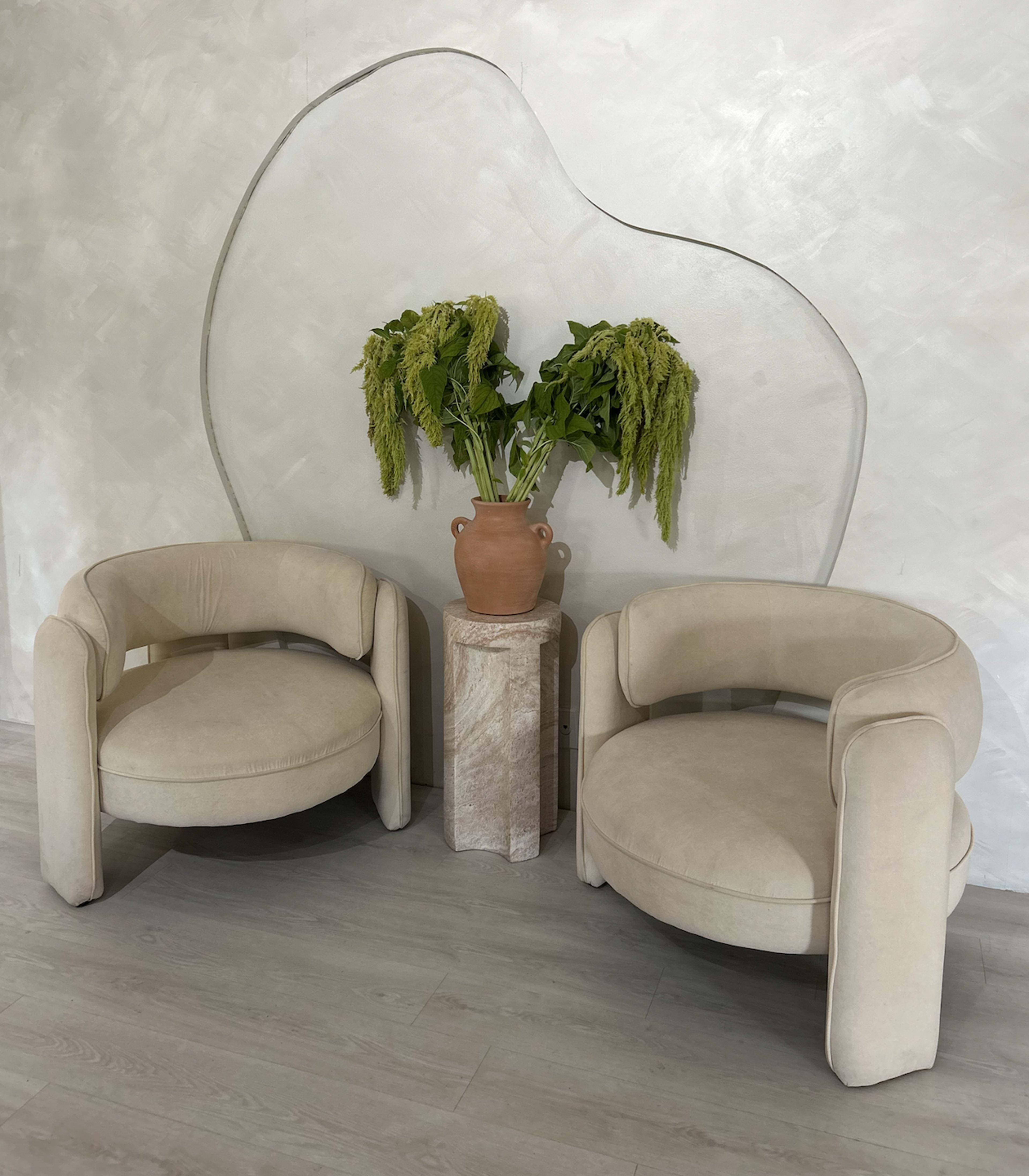 The image shows two rounded, beige chairs positioned beside a pedestal with a terracotta vase containing green plants, set against a textured wall.