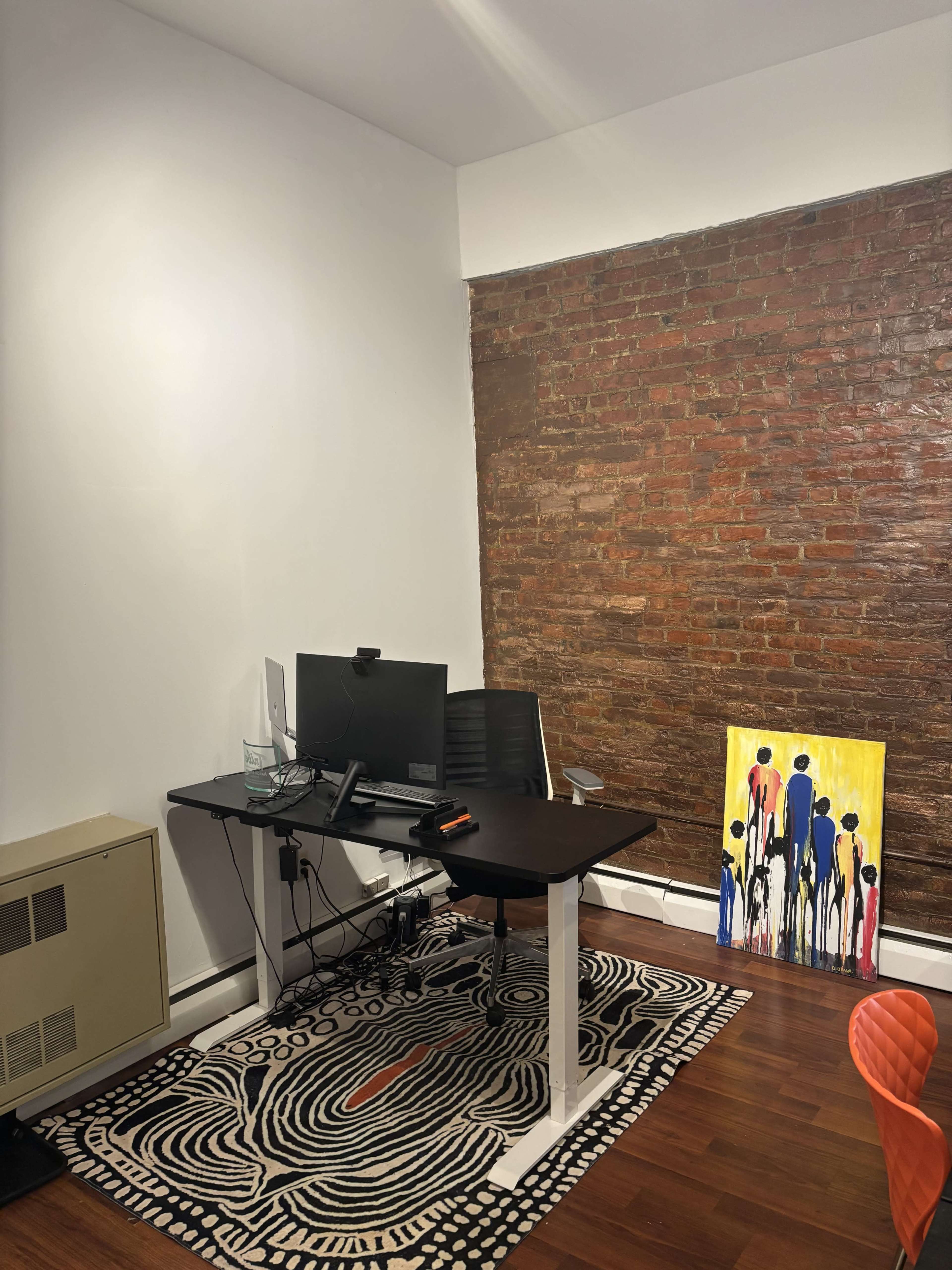 The image shows a minimalist office setup with a desk, computer, and a colorful painting against a brick wall, accompanied by a patterned rug and an orange chair.