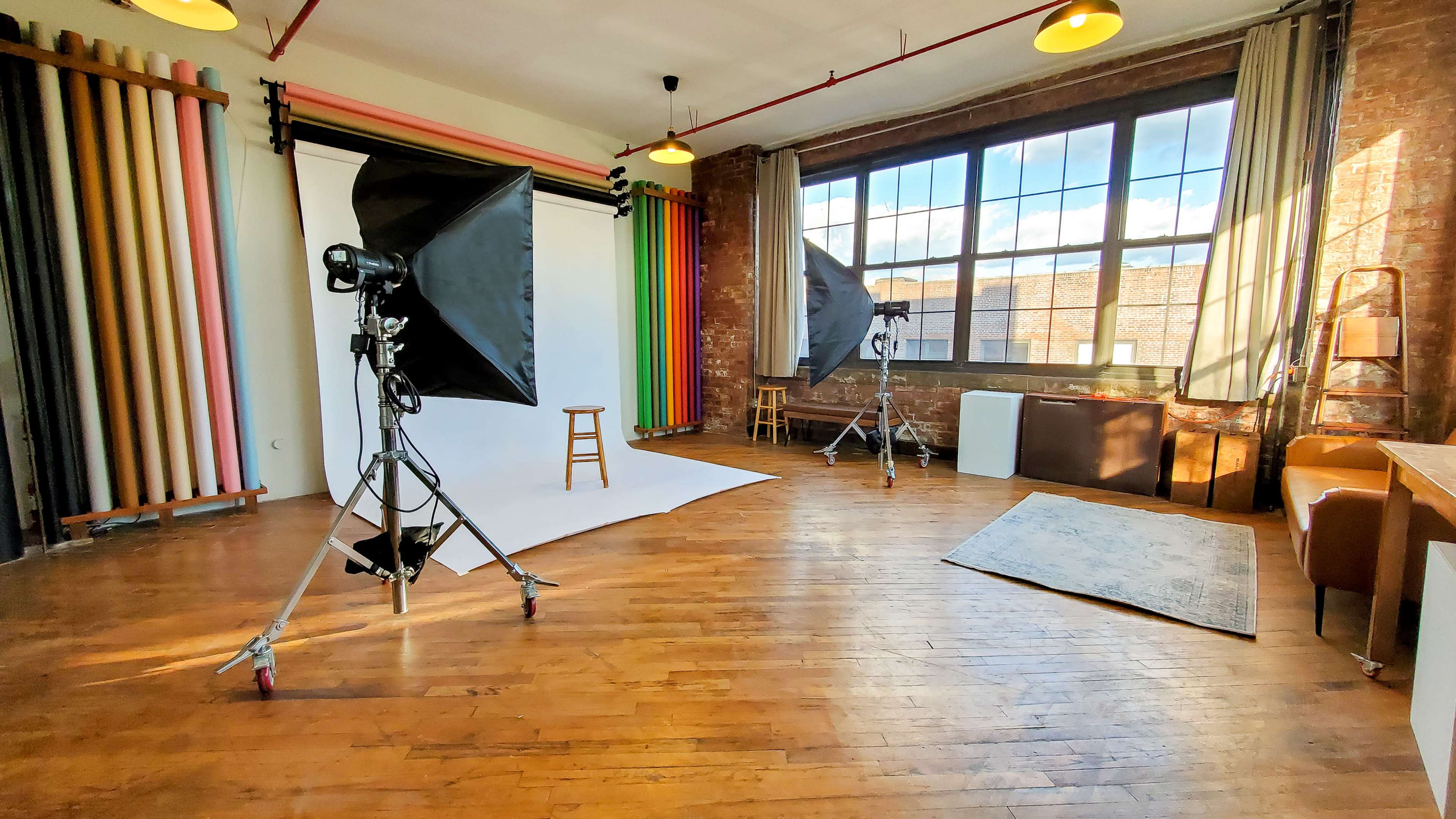 A photography studio with a white backdrop, lighting equipment, a stool, and a large window illuminated by natural light.