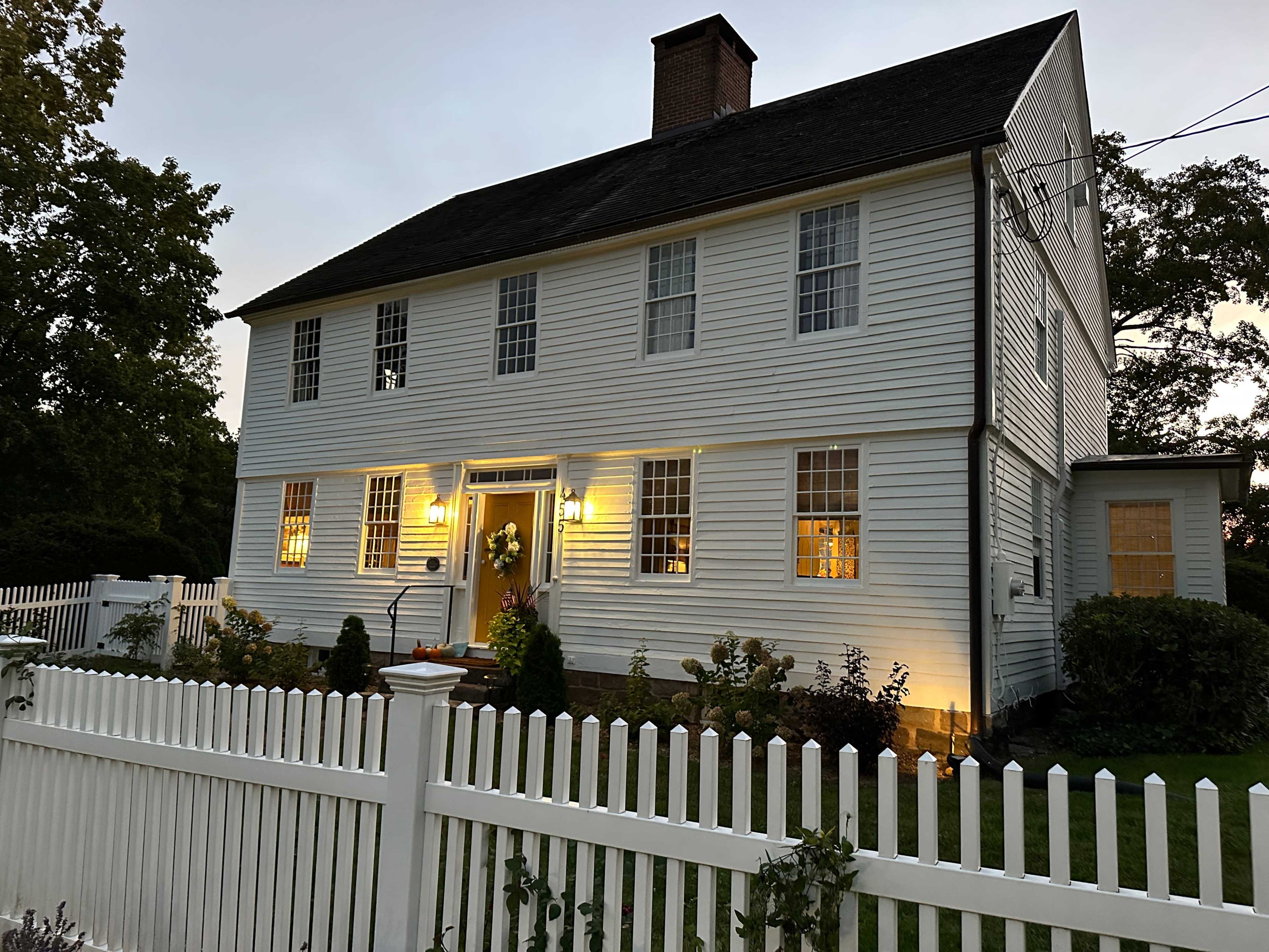 A two-story white house with a sloped roof and front porch is illuminated by warm lights against a dusk sky, surrounded by a white picket fence and landscaped greenery.