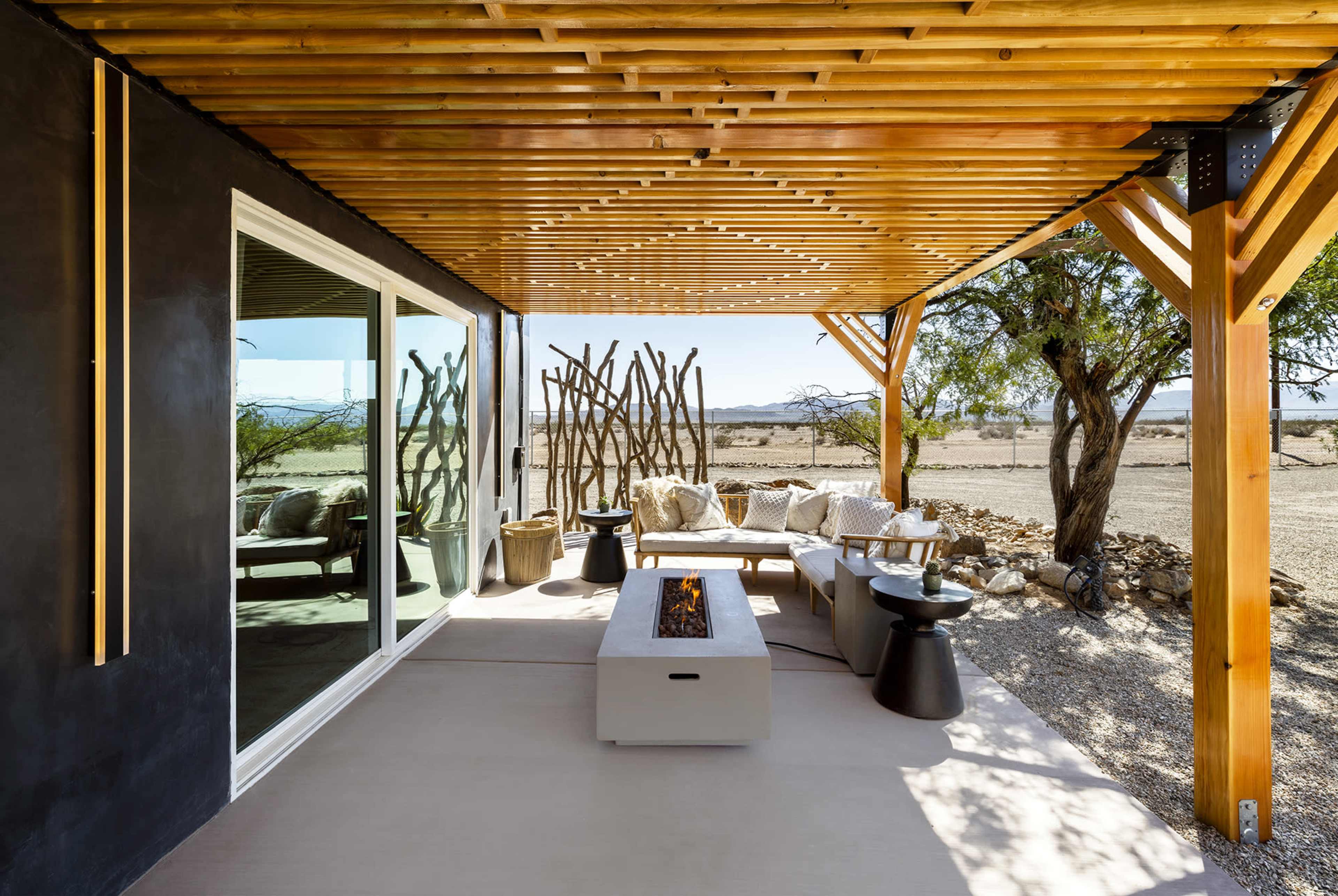 A modern outdoor seating area with a fire pit and a wooden pergola, overlooking a desert landscape.