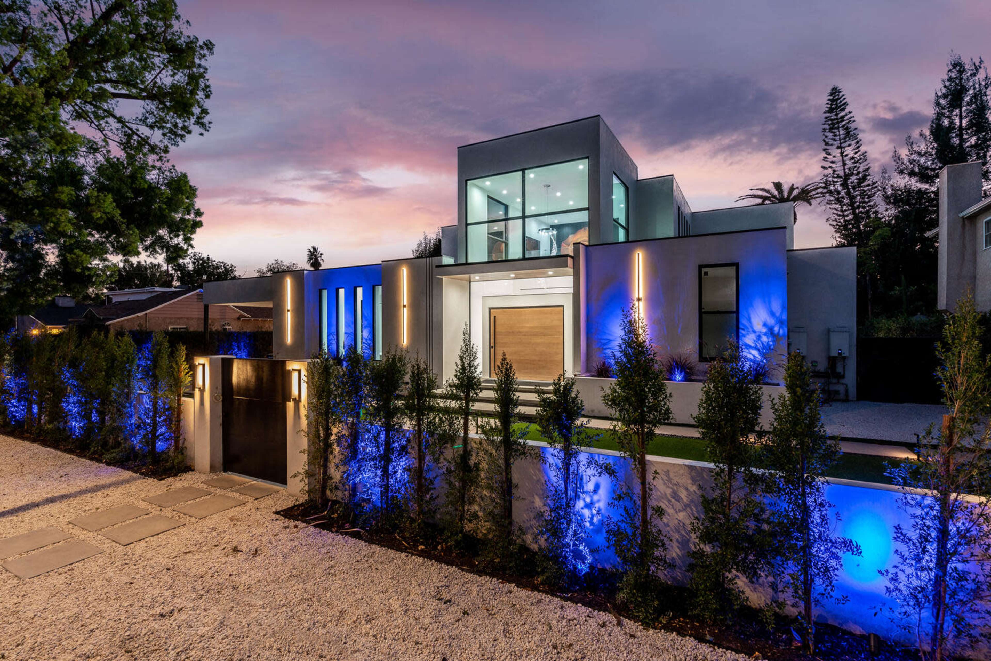 A modern two-story house with large glass windows, illuminated by blue lighting along the front pathway and landscaping, set against a sunset sky.