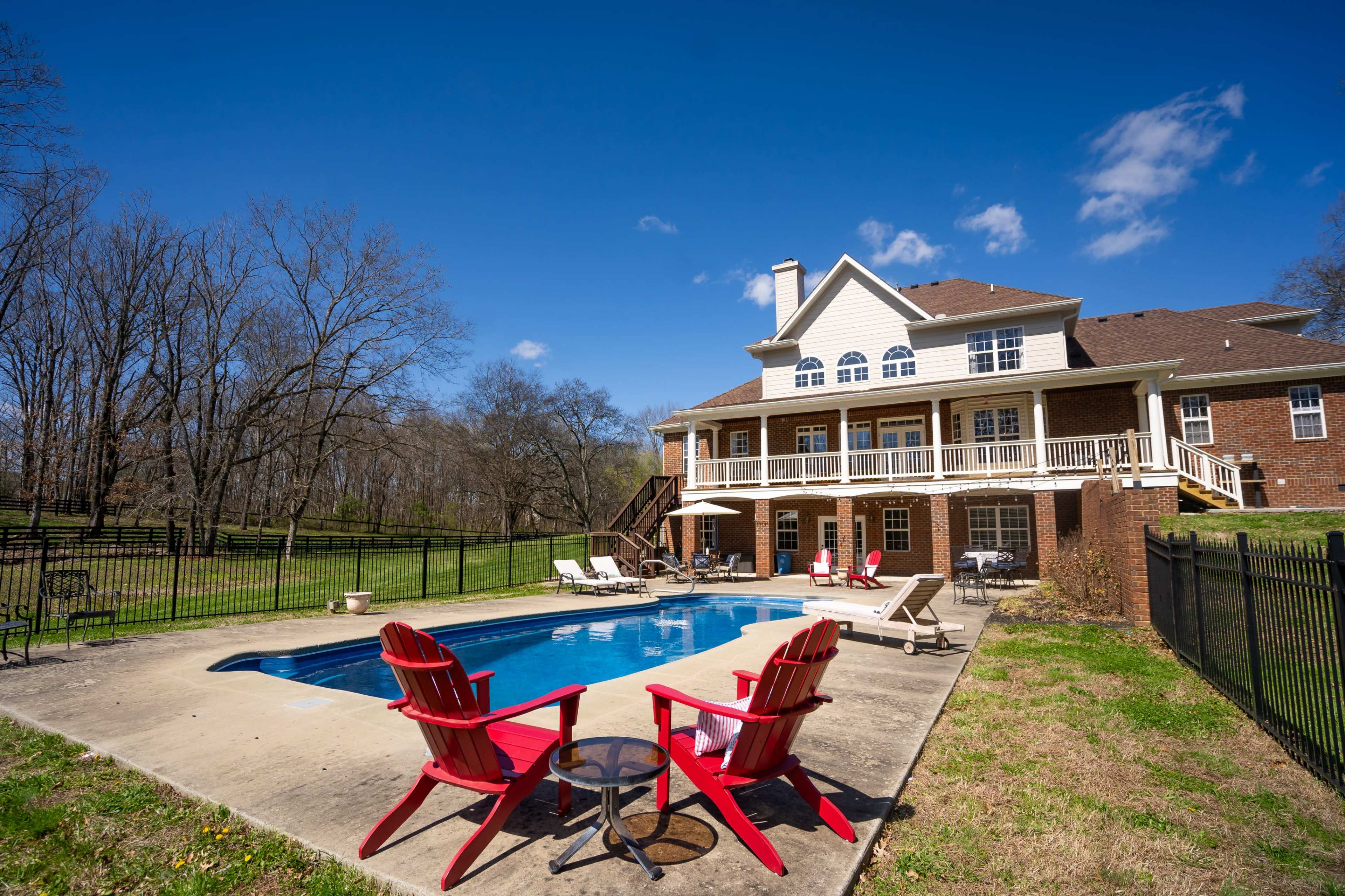 A backyard features a swimming pool surrounded by lounge chairs and a patio, with a spacious two-story house in the background.