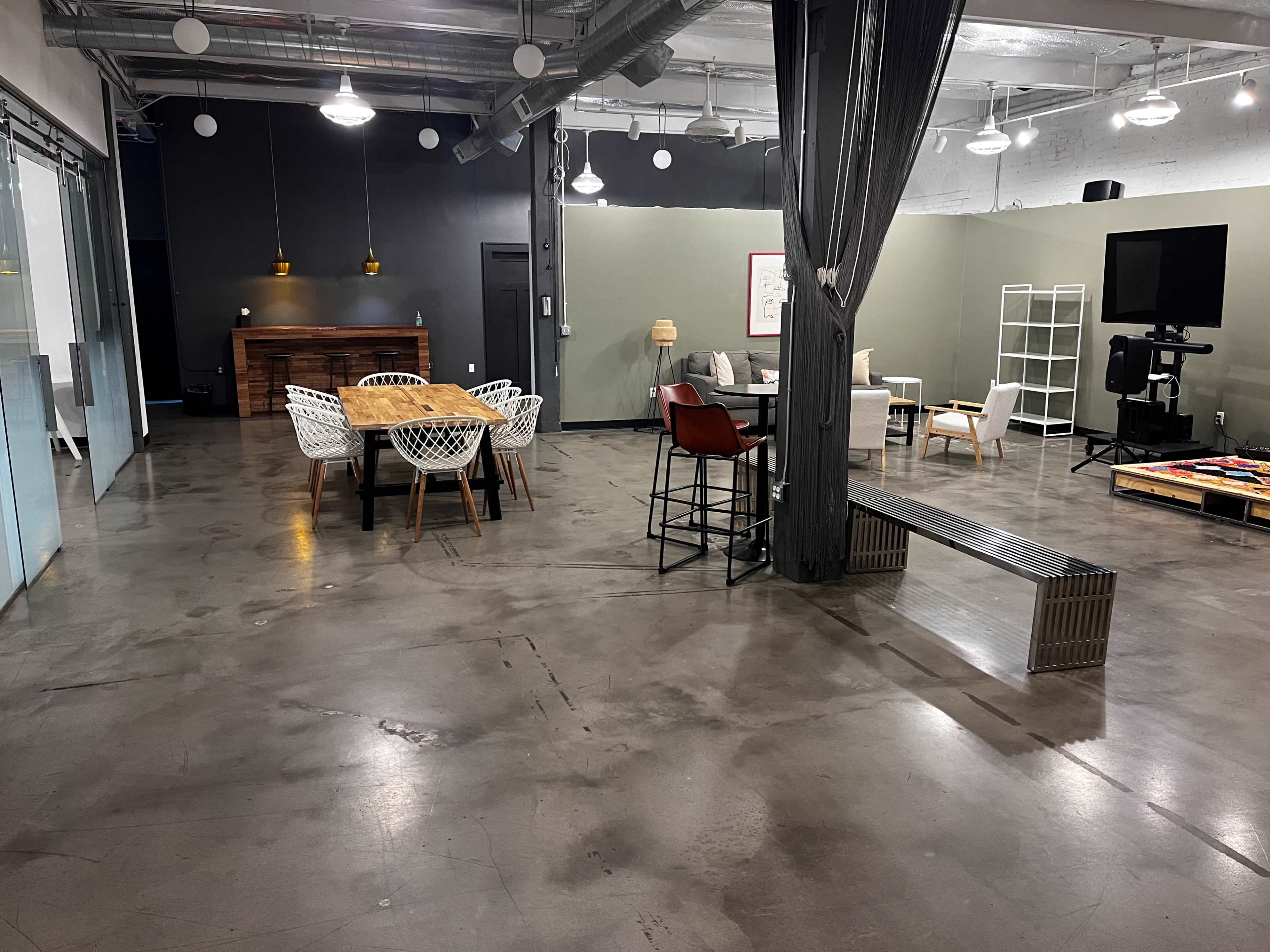 The image shows a spacious, modern office interior with various seating arrangements, a wooden dining table, and a lounge area.