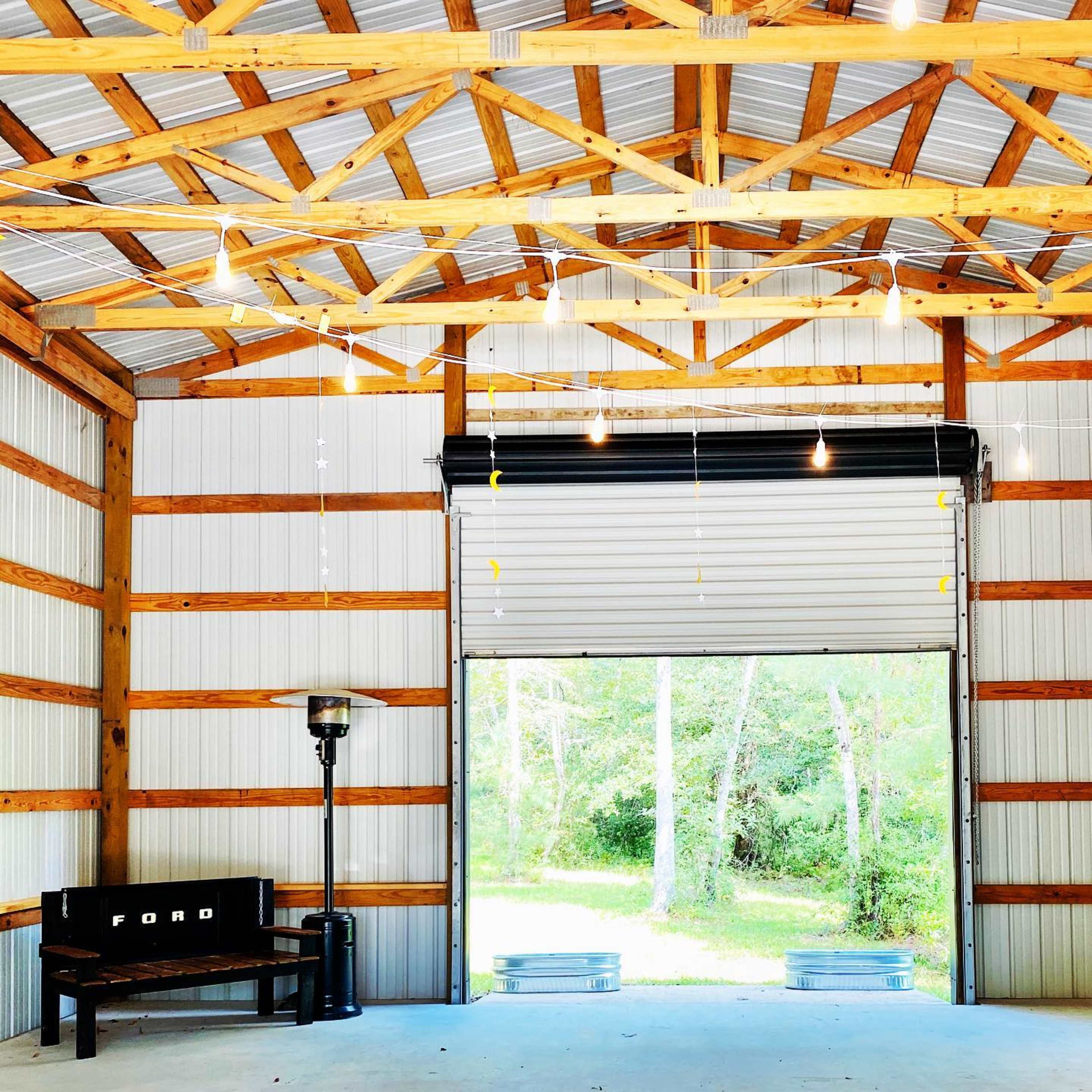 The interior of a wooden barn with a large open garage door, a bench, and a light fixture, set against a backdrop of trees.