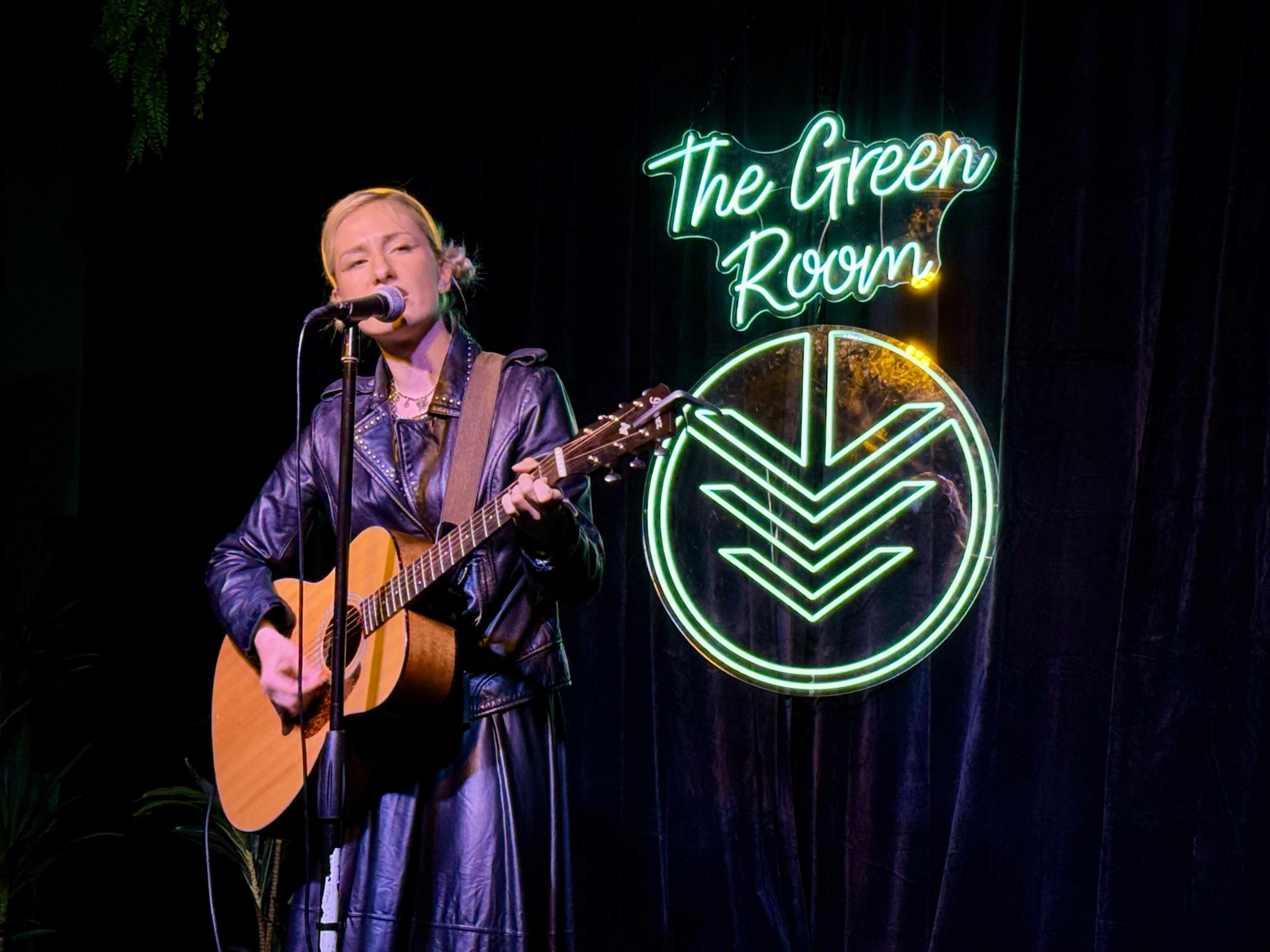 A woman in a black leather jacket plays an acoustic guitar and sings on stage in front of a neon sign that reads "The Green Room."