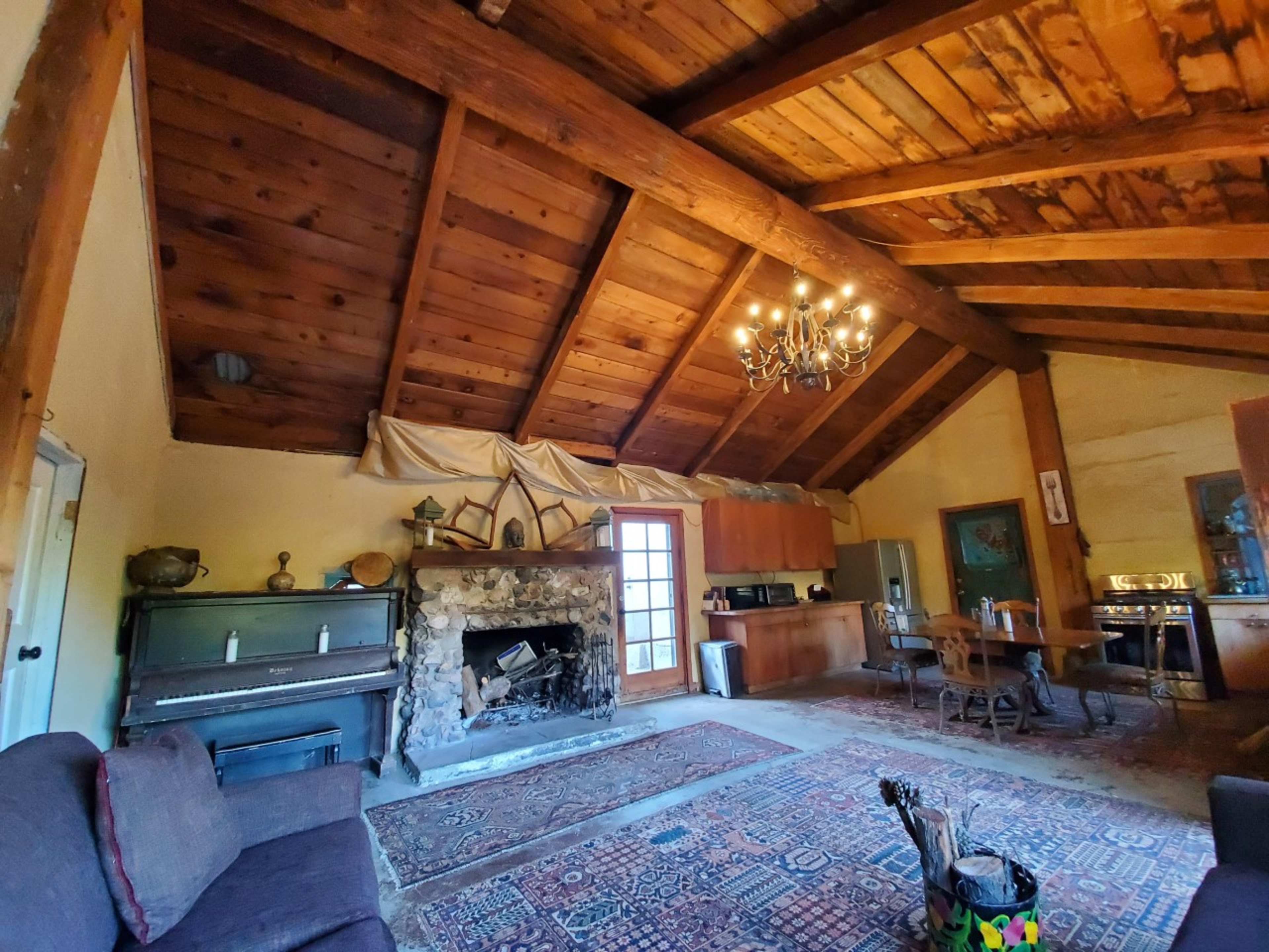 A rustic living room with a wooden beamed ceiling, a stone fireplace, a piano, and various pieces of furniture.