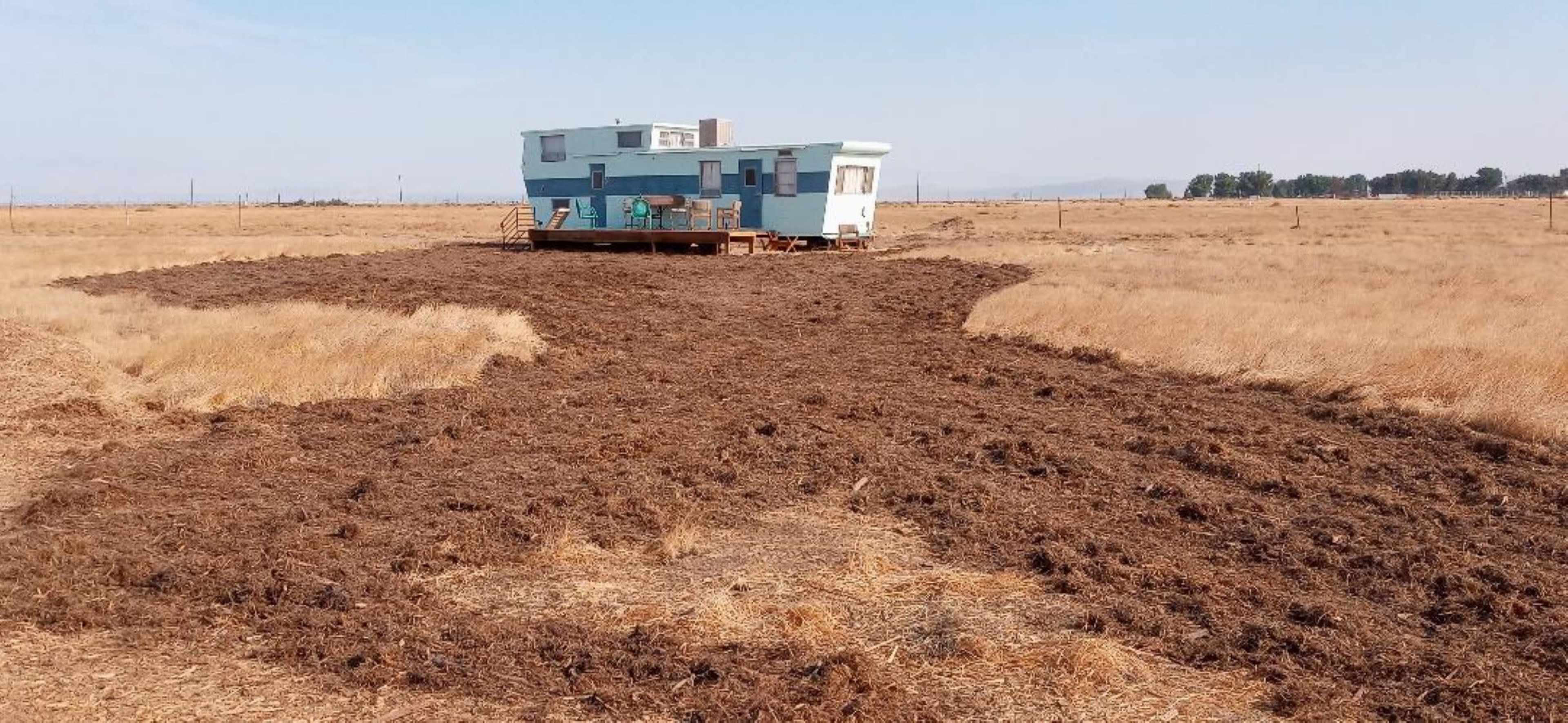 A blue trailer sits on a patch of dry grass surrounded by a large area of freshly turned soil.
