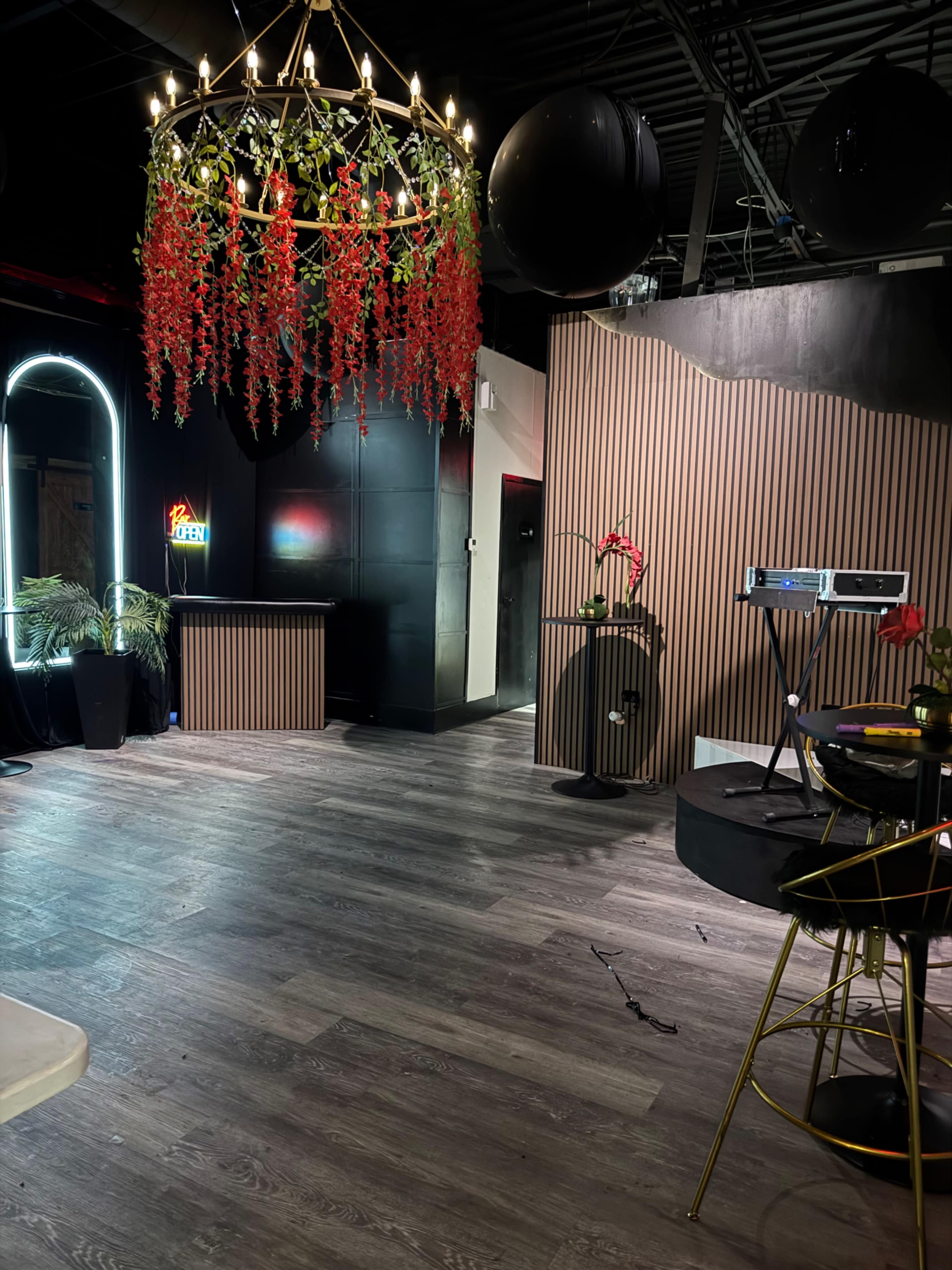 The image shows a stylish interior space featuring a chandelier with red flowers, black walls, and a wooden floor, along with various decorative elements like a projector and plants.