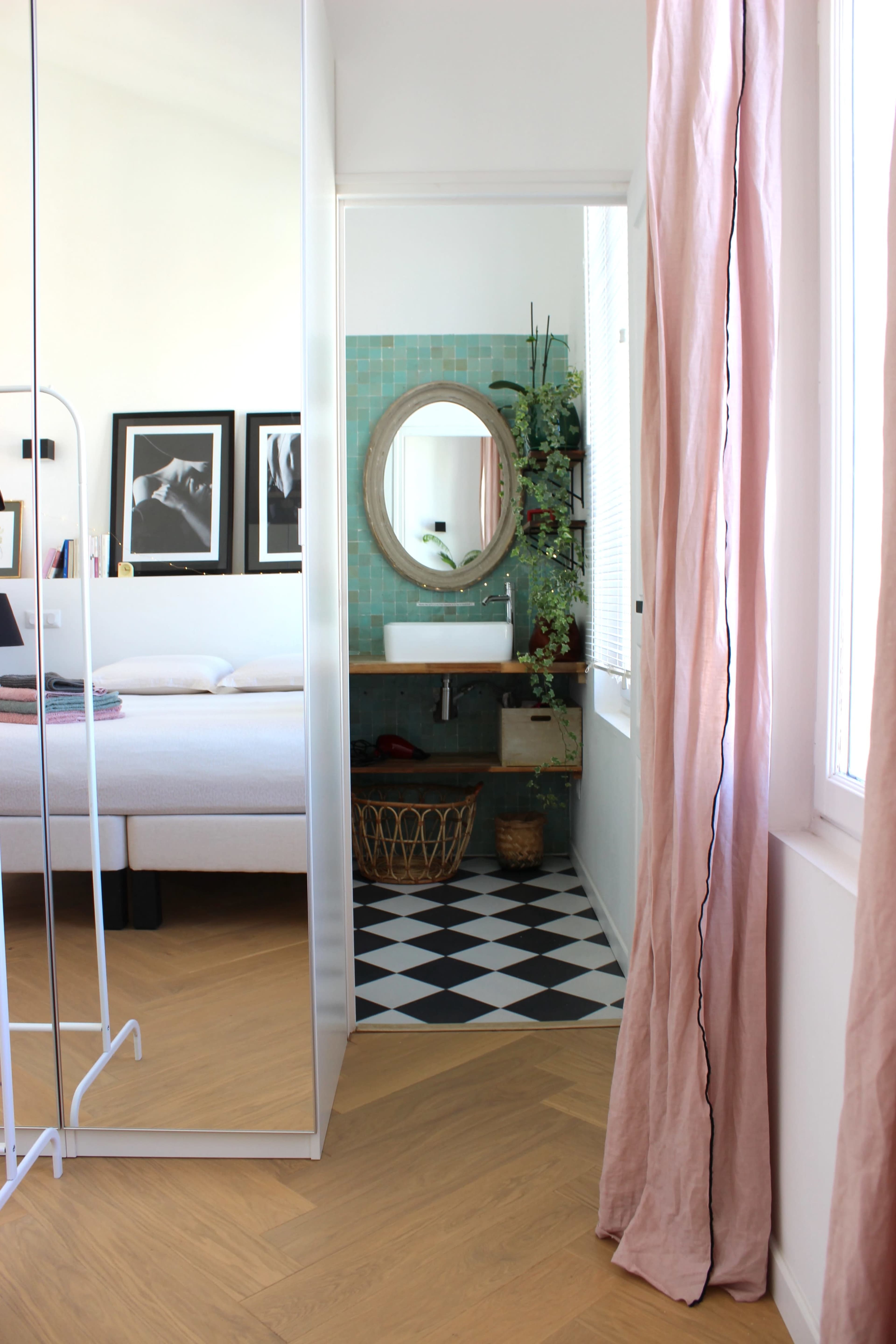 A bright room features a large mirror reflecting a bathroom with a round mirror, green tiles, and a patterned floor, separated by a doorway.