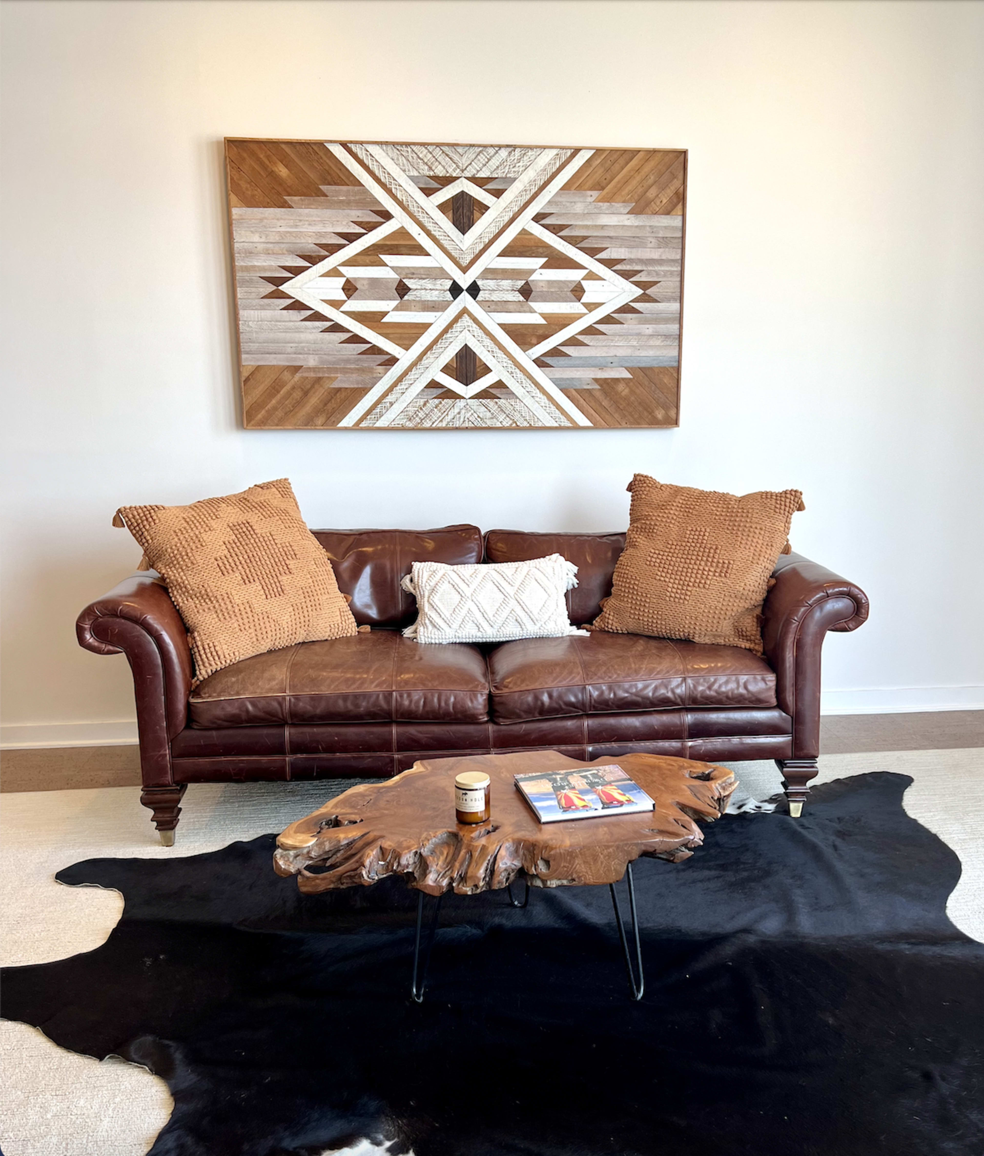 A brown leather sofa with textured pillows sits in front of a wooden wall art piece, accompanied by a coffee table made from a polished wood slab on a black and white cowhide rug.