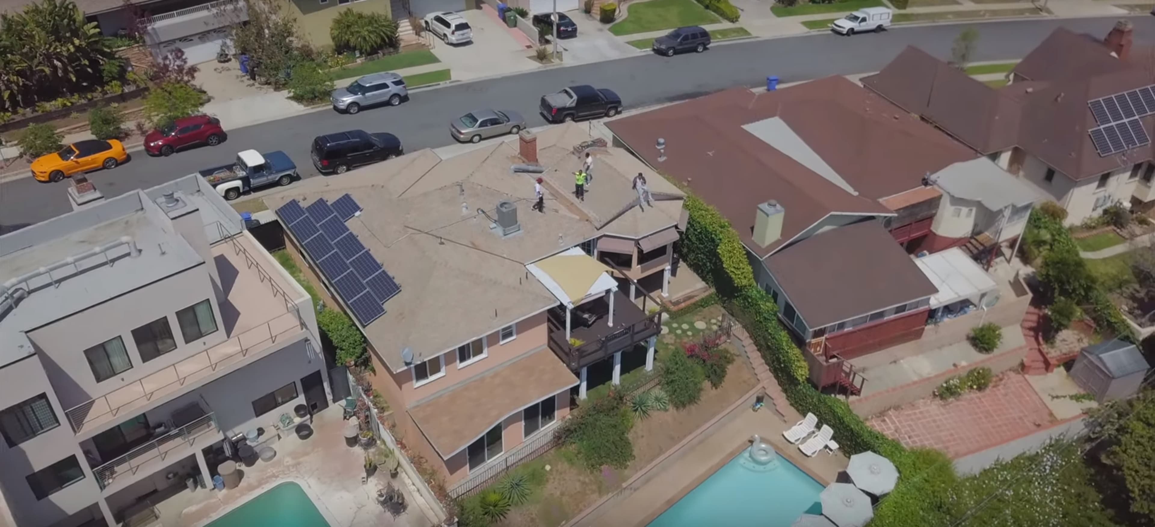 An aerial view shows workers on the roof of a house with solar panels, alongside several parked cars and a swimming pool in the backyard.