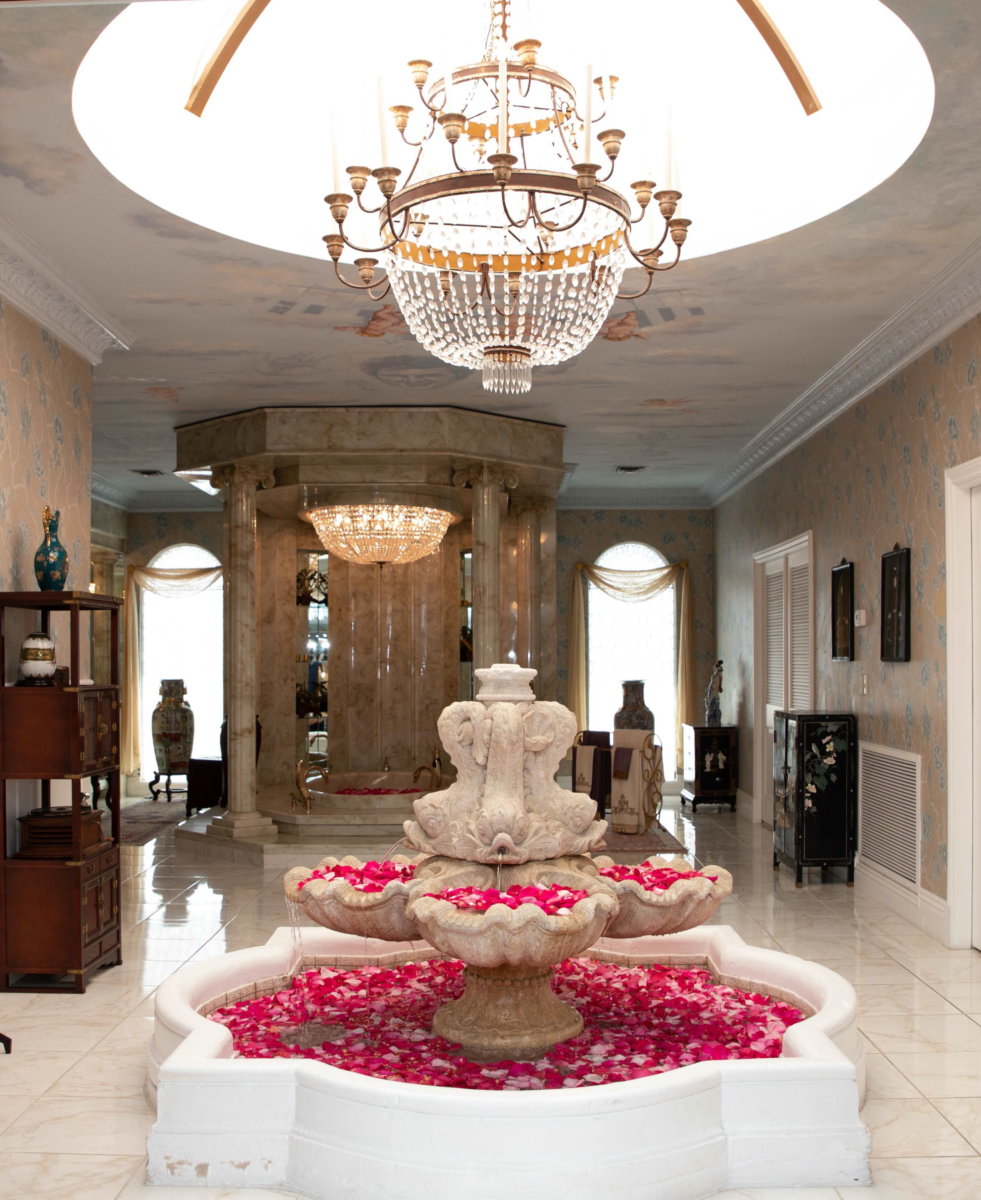 A spacious entryway featuring a decorative fountain surrounded by rose petals, with a chandelier overhead and elegant furnishings against ornate wallpaper.