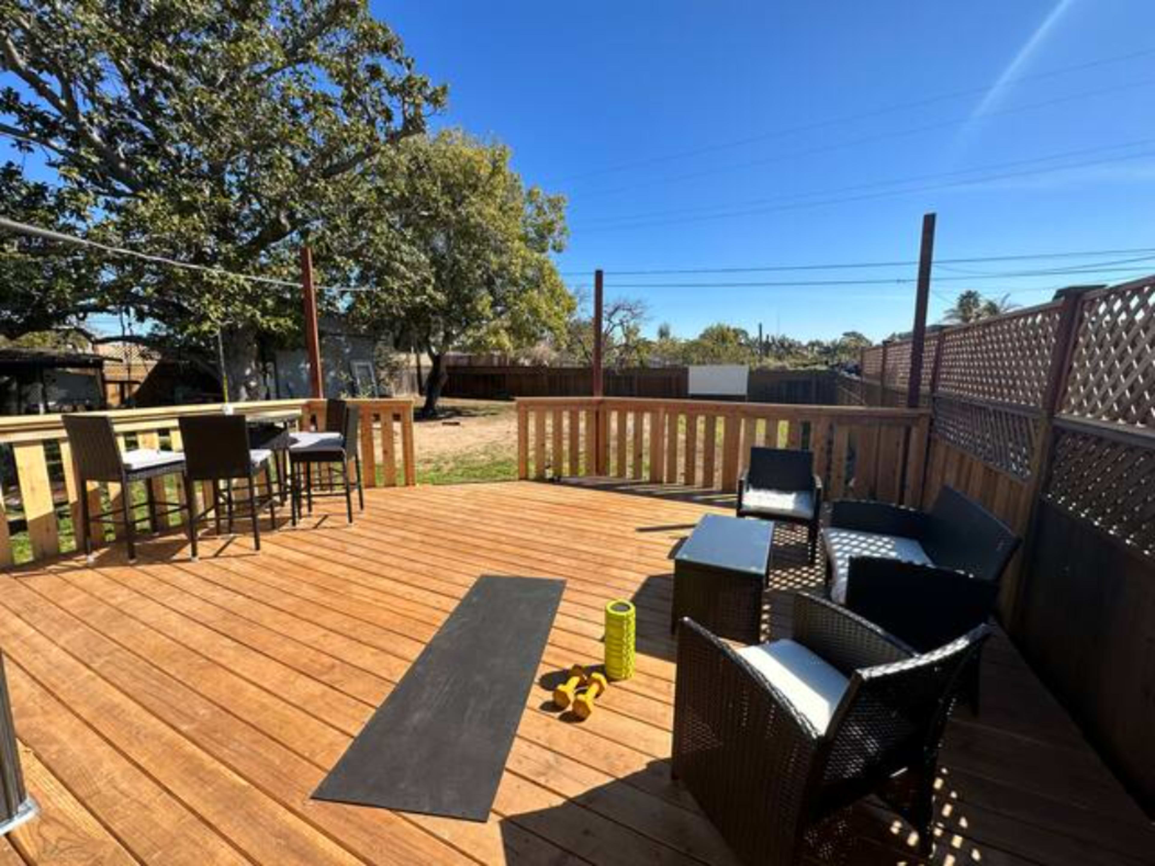 The image shows a wooden deck featuring outdoor seating, a yoga mat, and a set of dumbbells, with clear skies and trees in the background.