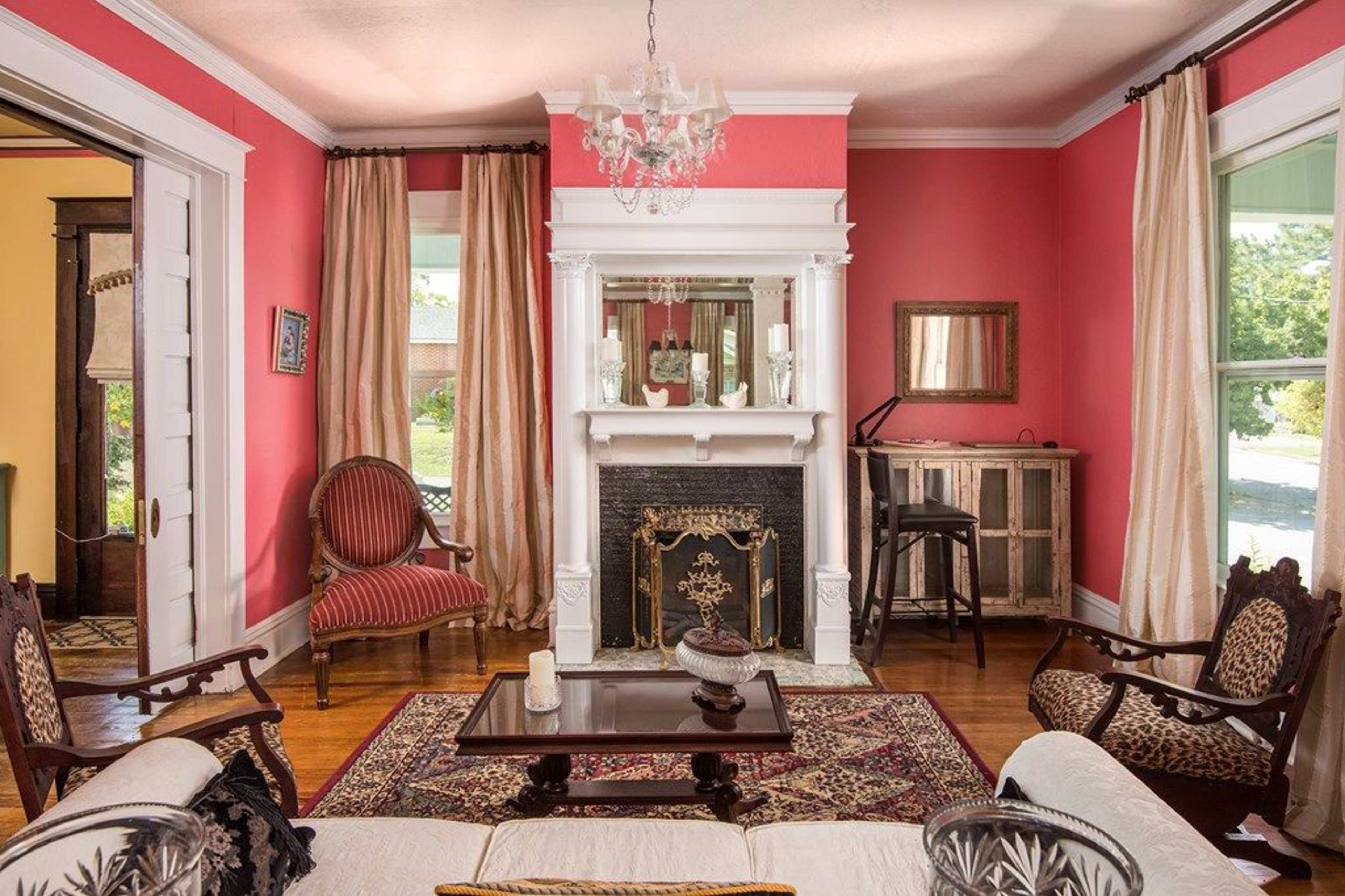 The room features a pink wall, a fireplace with a mirror above it, two armchairs, and a small table across from a sofa, all illuminated by natural light.