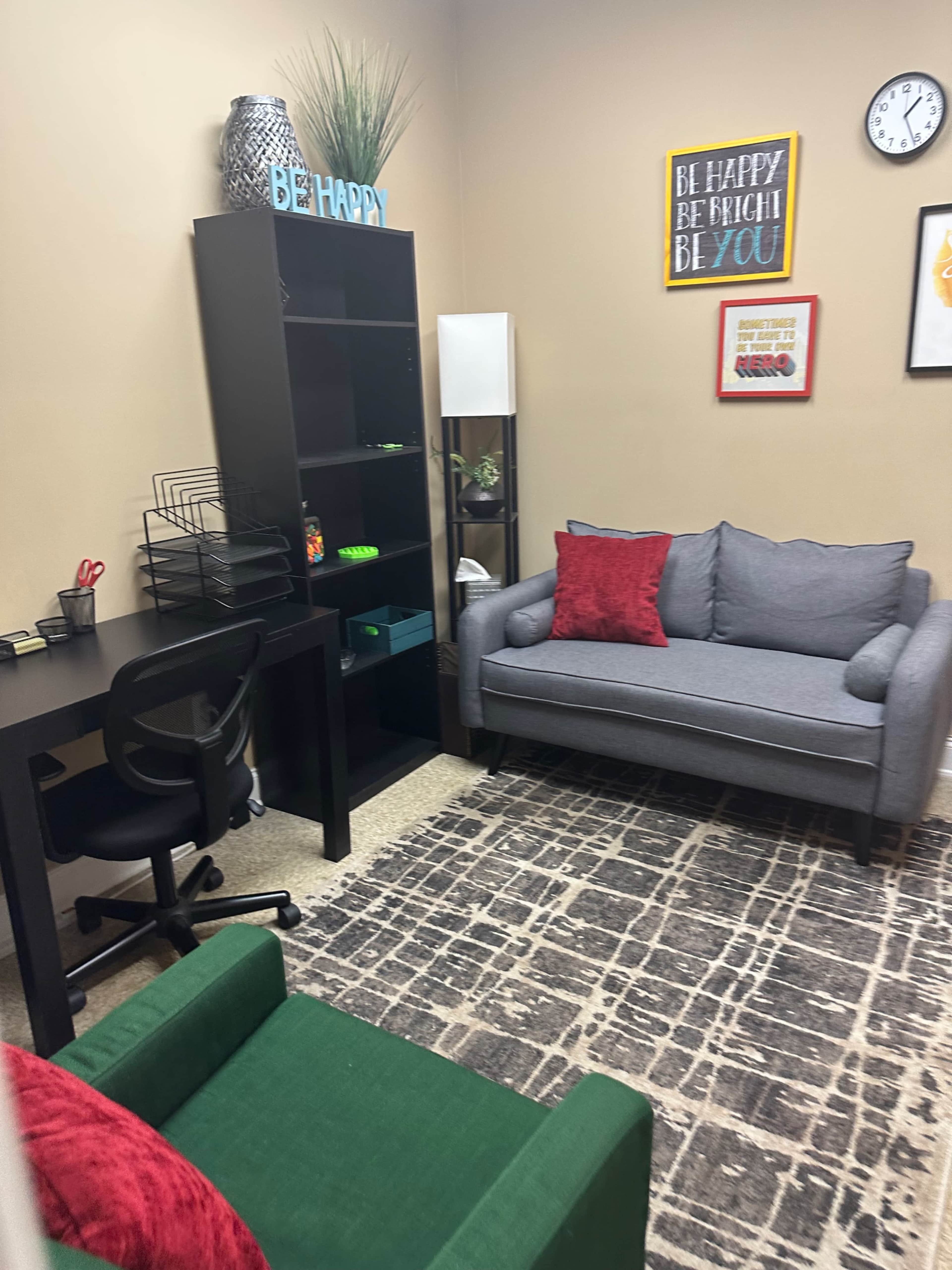 The image shows a small office space featuring a gray couch, a green chair, a black desk with a rolling chair, a black bookshelf, and decorations on the walls.