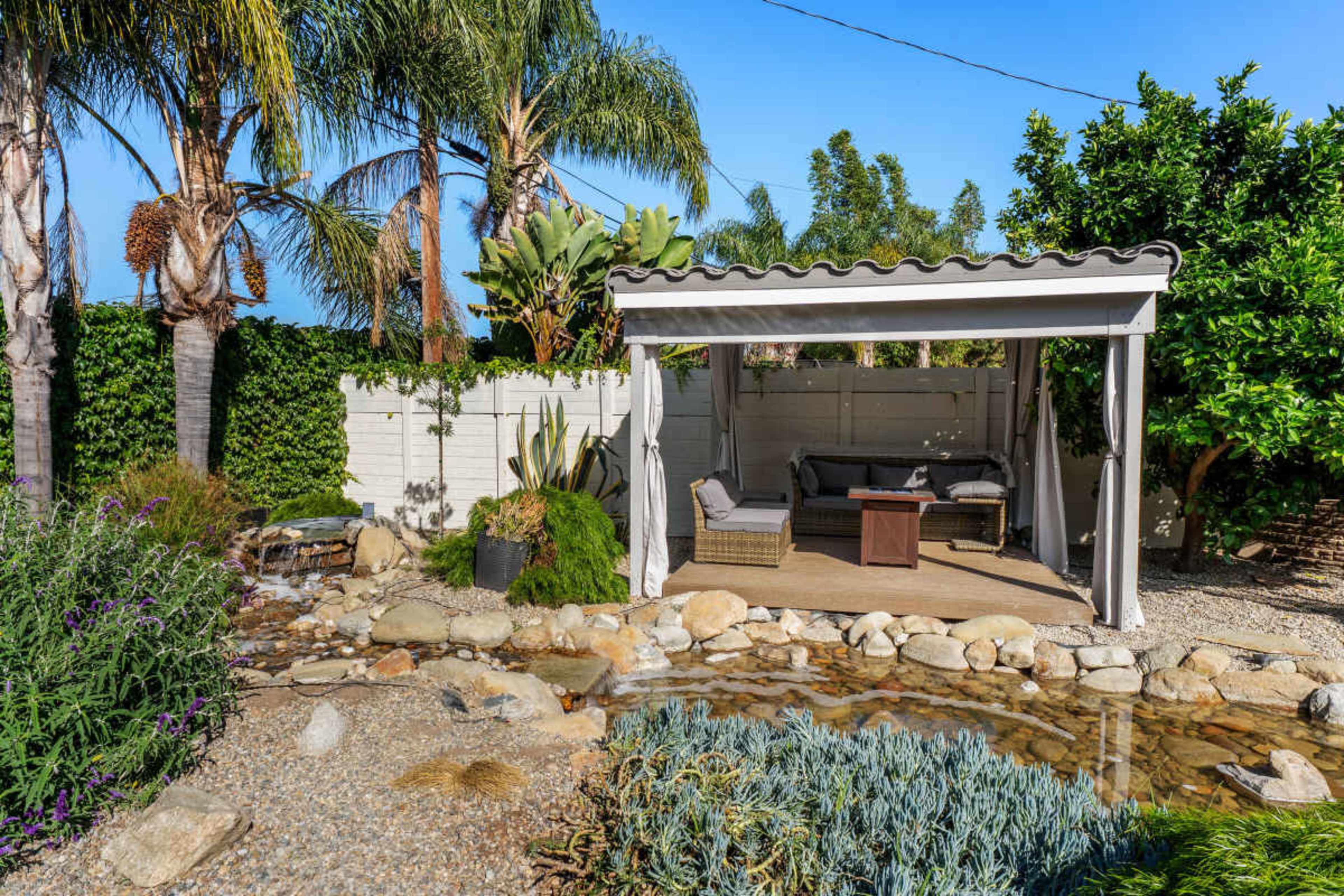 A shaded outdoor seating area sits among palm trees and lush plants, next to a decorative stream and rocky landscape.