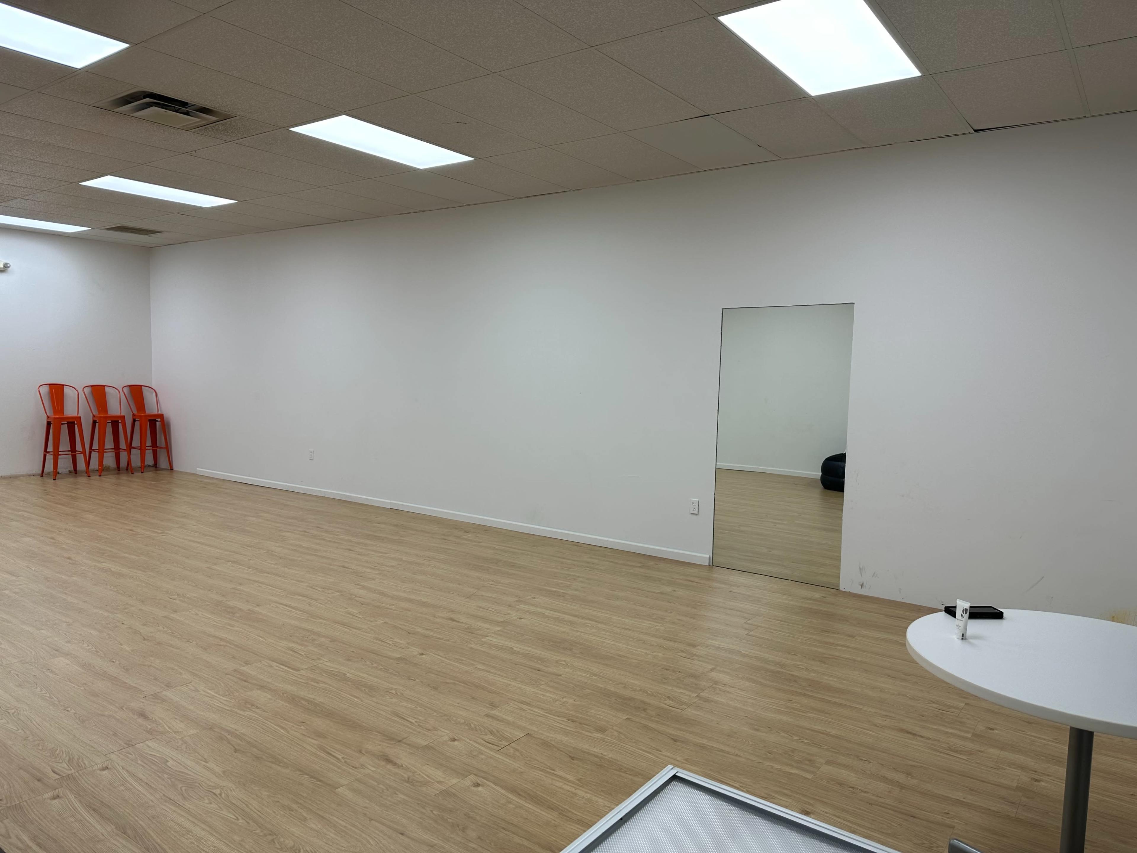 The image shows a spacious room with light wooden flooring, a blank white wall, a large mirror, a round table with a pen, and four orange chairs stacked against another wall.