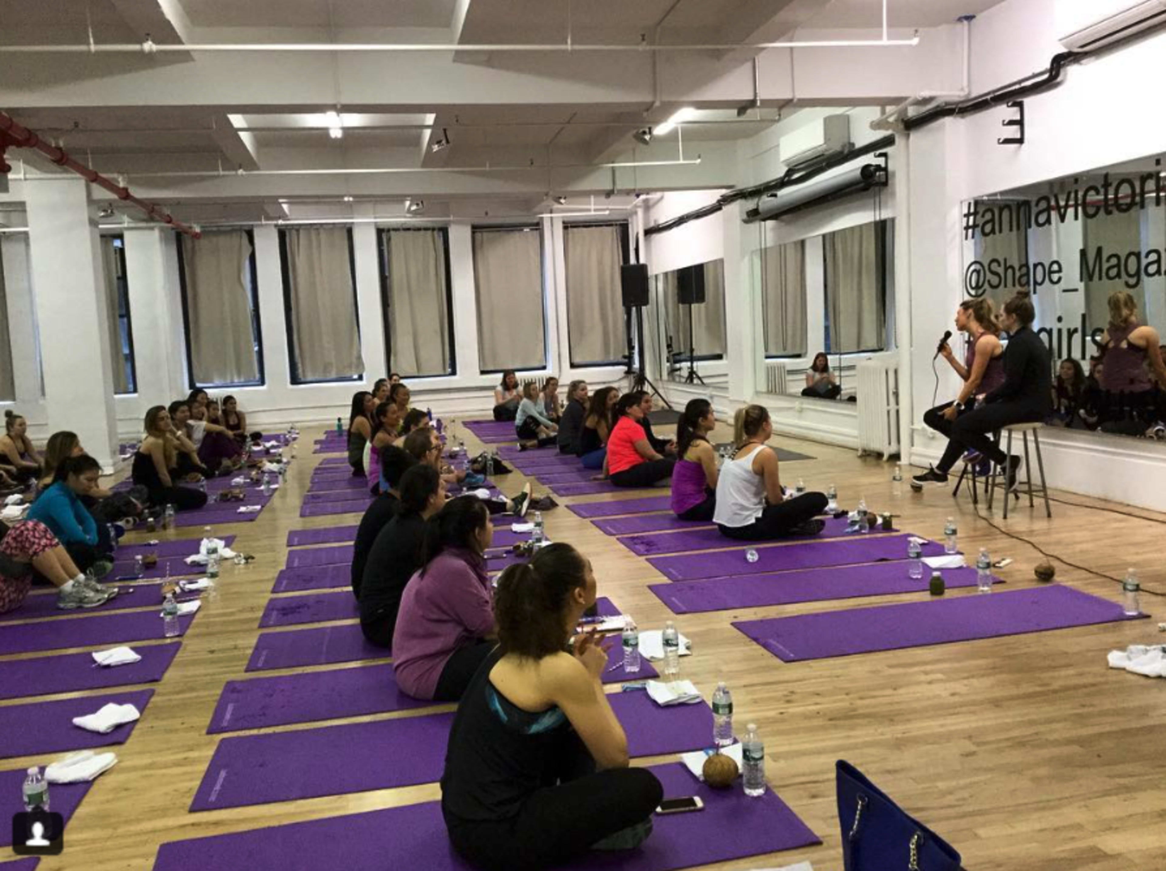 A group of women participates in a yoga class on purple mats in a well-lit studio.