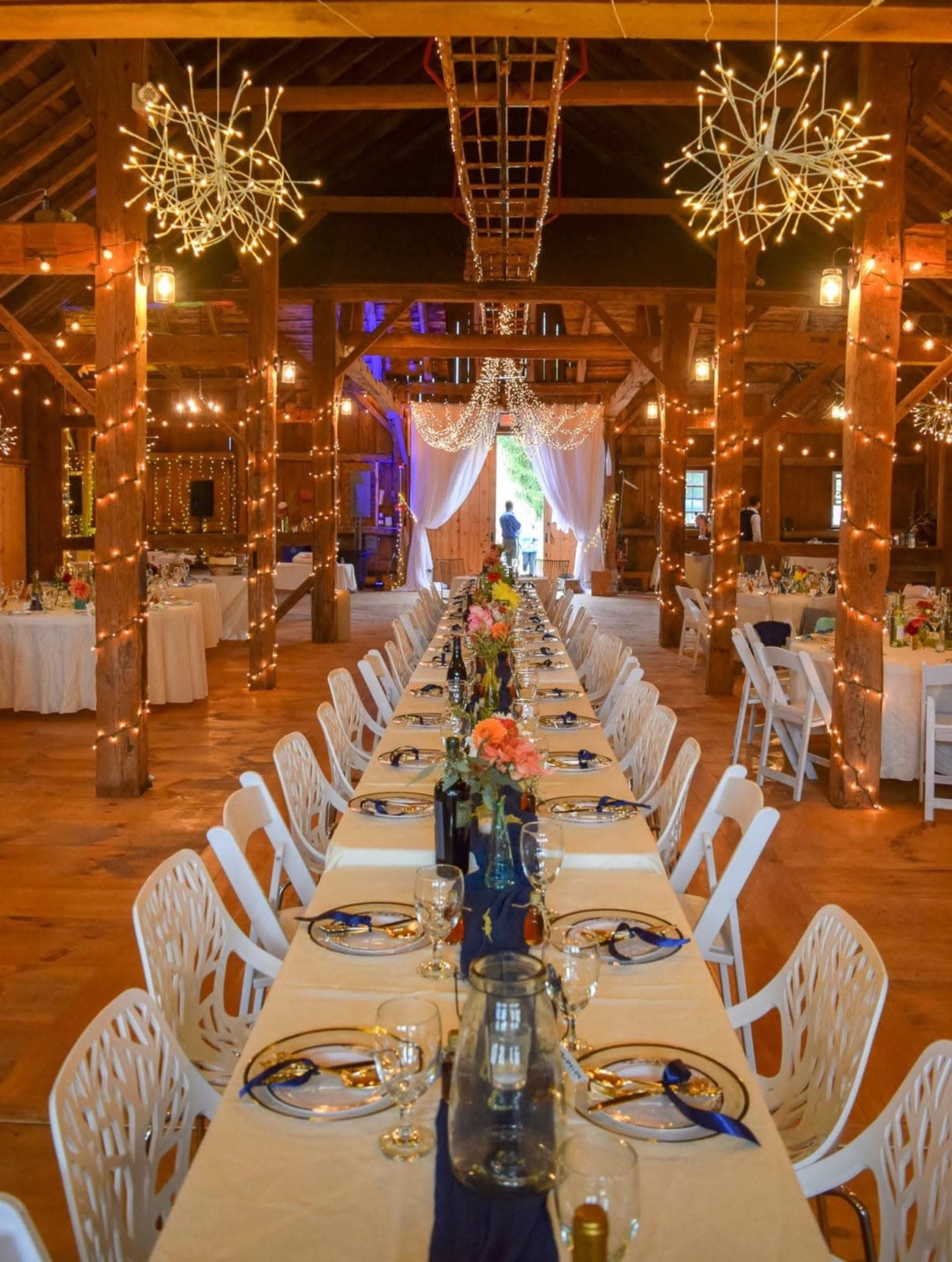 A long banquet table is elegantly set up in a spacious barn, decorated with fairy lights and floral arrangements, leading to a backdrop of draped fabric.