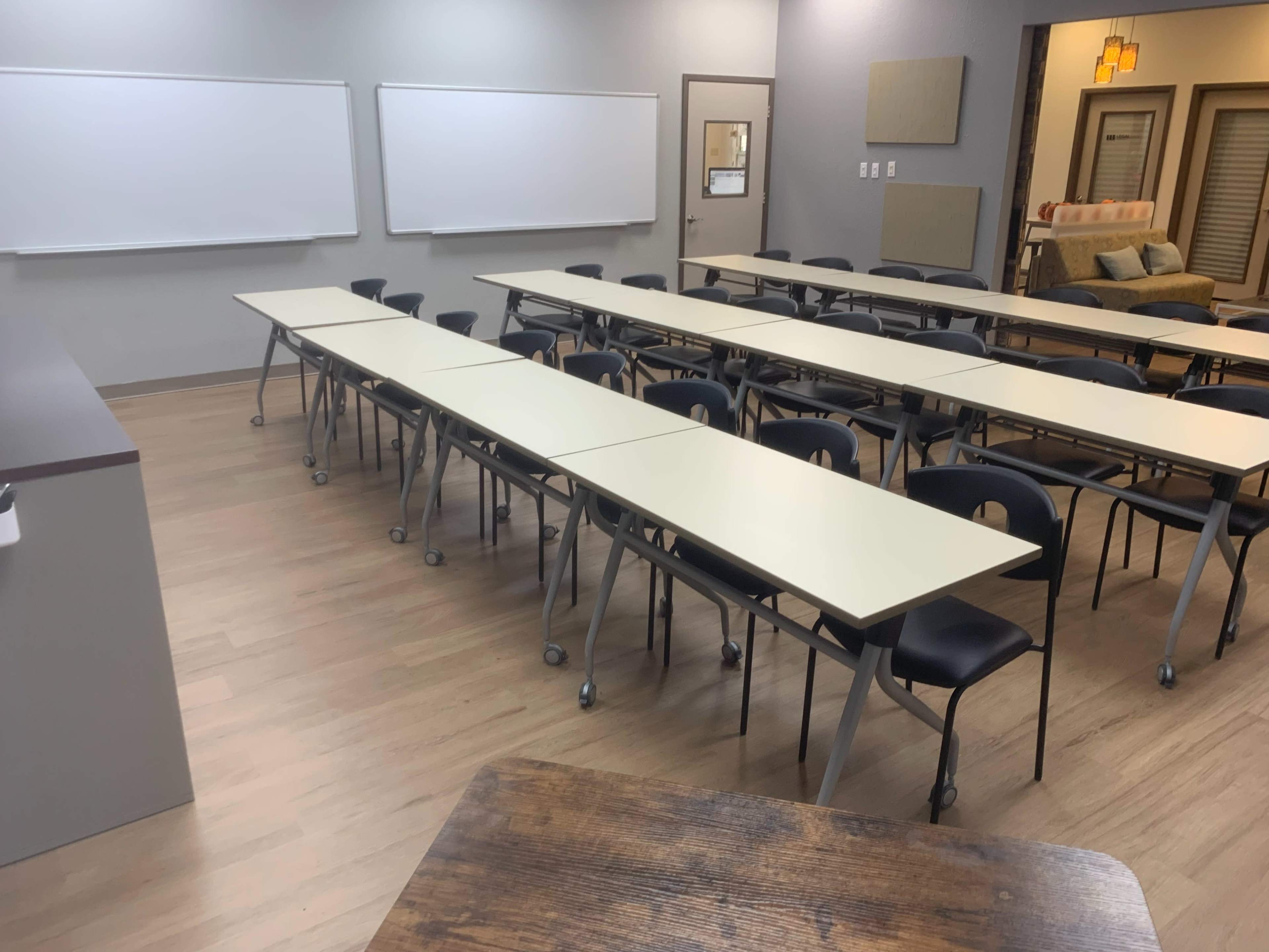 A classroom with multiple rows of tables and chairs facing a front area equipped with whiteboards.