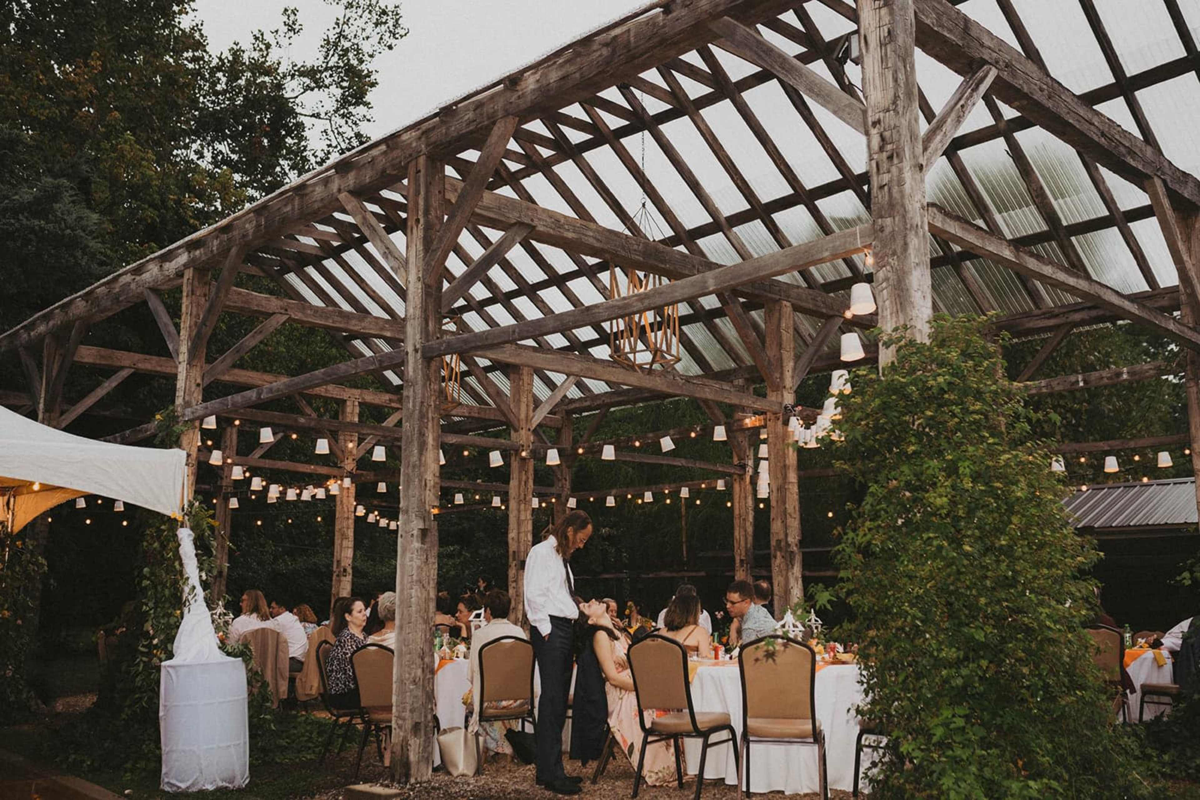 A wooden structure with a slatted roof and overhead lights hosts a dinner gathering with guests seated at tables.