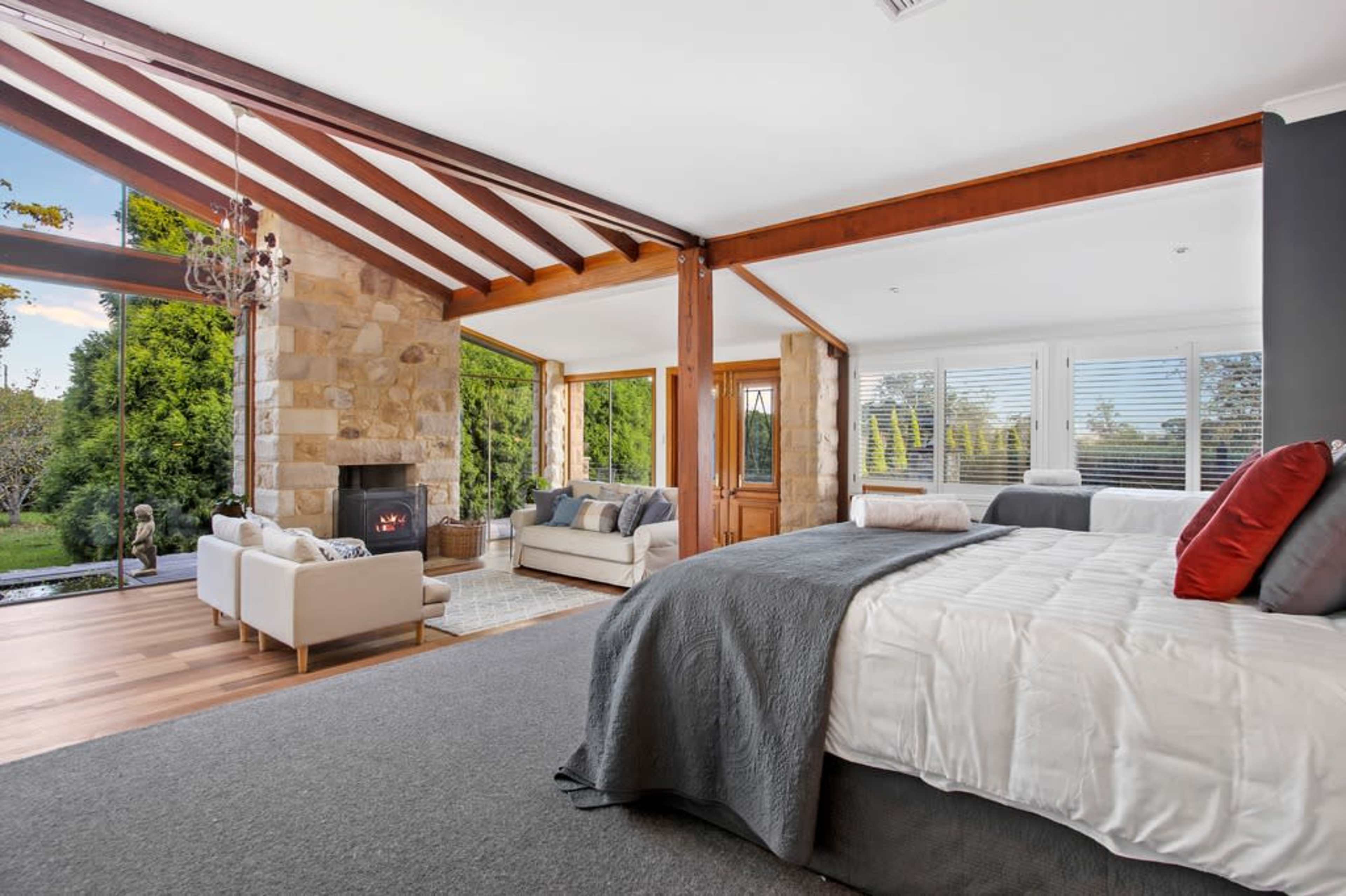 A spacious bedroom with a large bed, a stone fireplace, and a seating area featuring two sofas, all illuminated by natural light from large windows.
