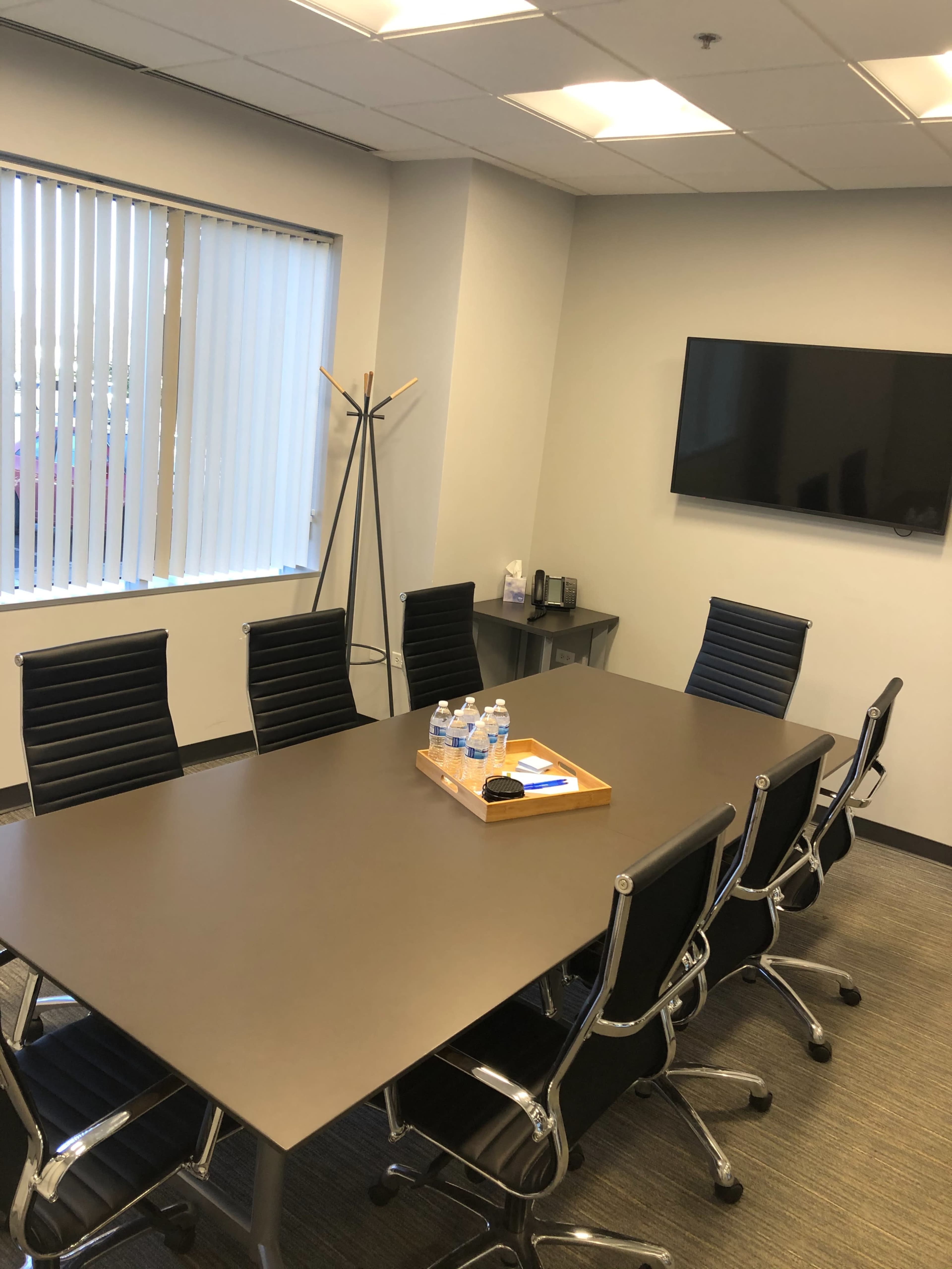 A modern conference room features a large table surrounded by ergonomic chairs, with a wall-mounted screen and a side table holding water bottles and stationery.